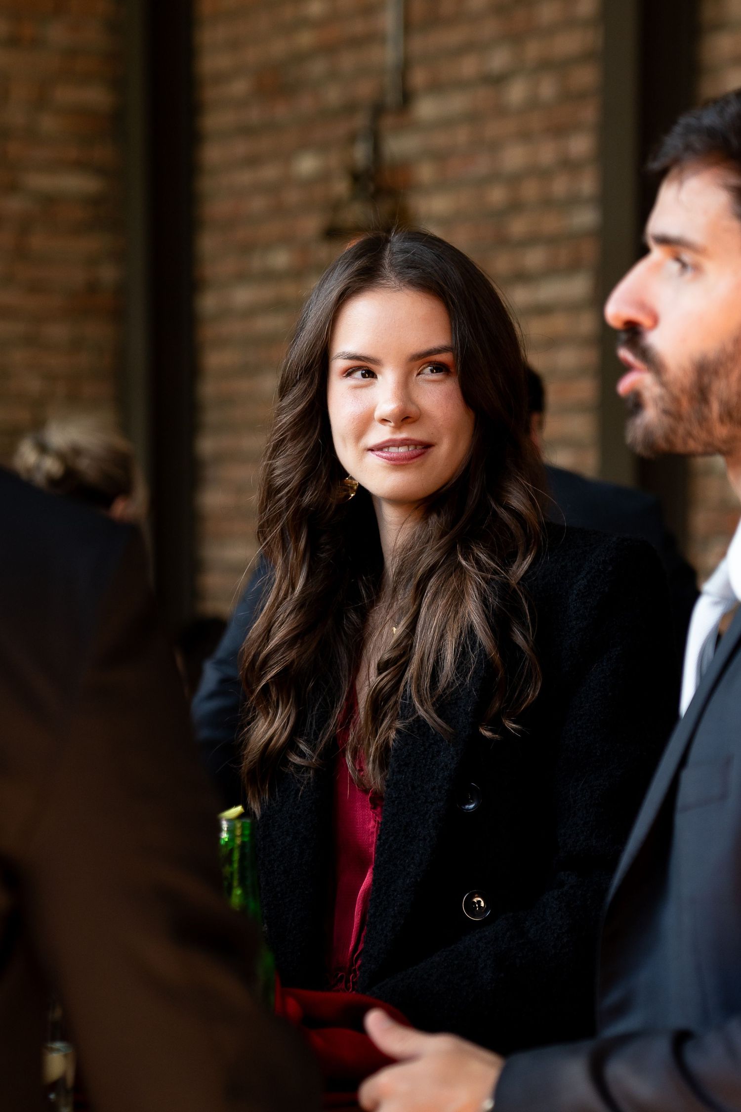 Young woman with long dark hair in black blazer and red top smiling at an indoor social gathering.