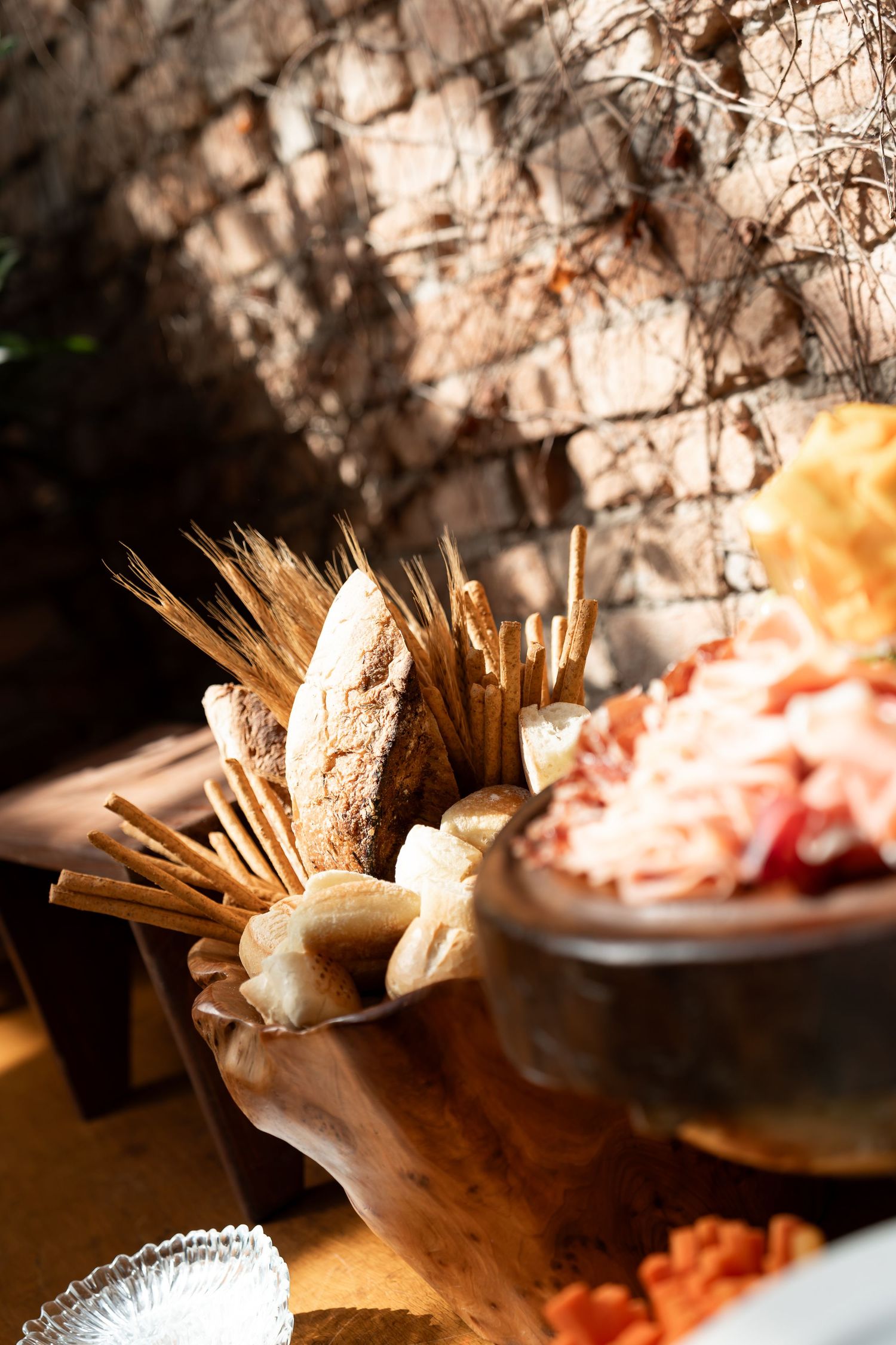 Rustic wooden board with artisan bread, breadsticks, and a bowl of charcuterie meats displayed outdoors in warm sunlight.