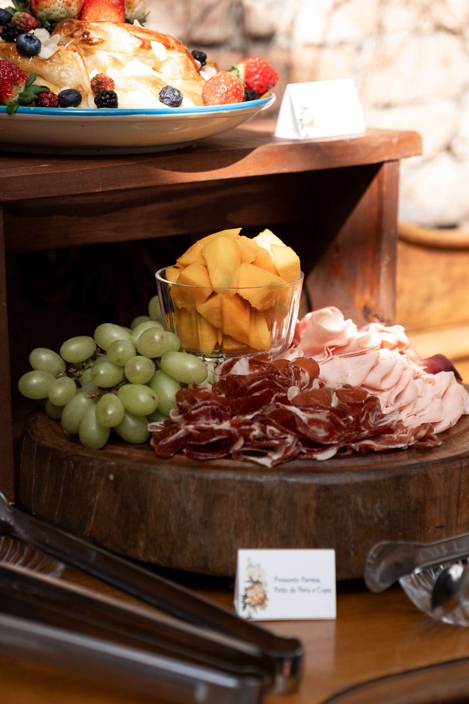 Charcuterie board with prosciutto, ham, green grapes, and melon cubes on wood, with a fruit dessert display above.