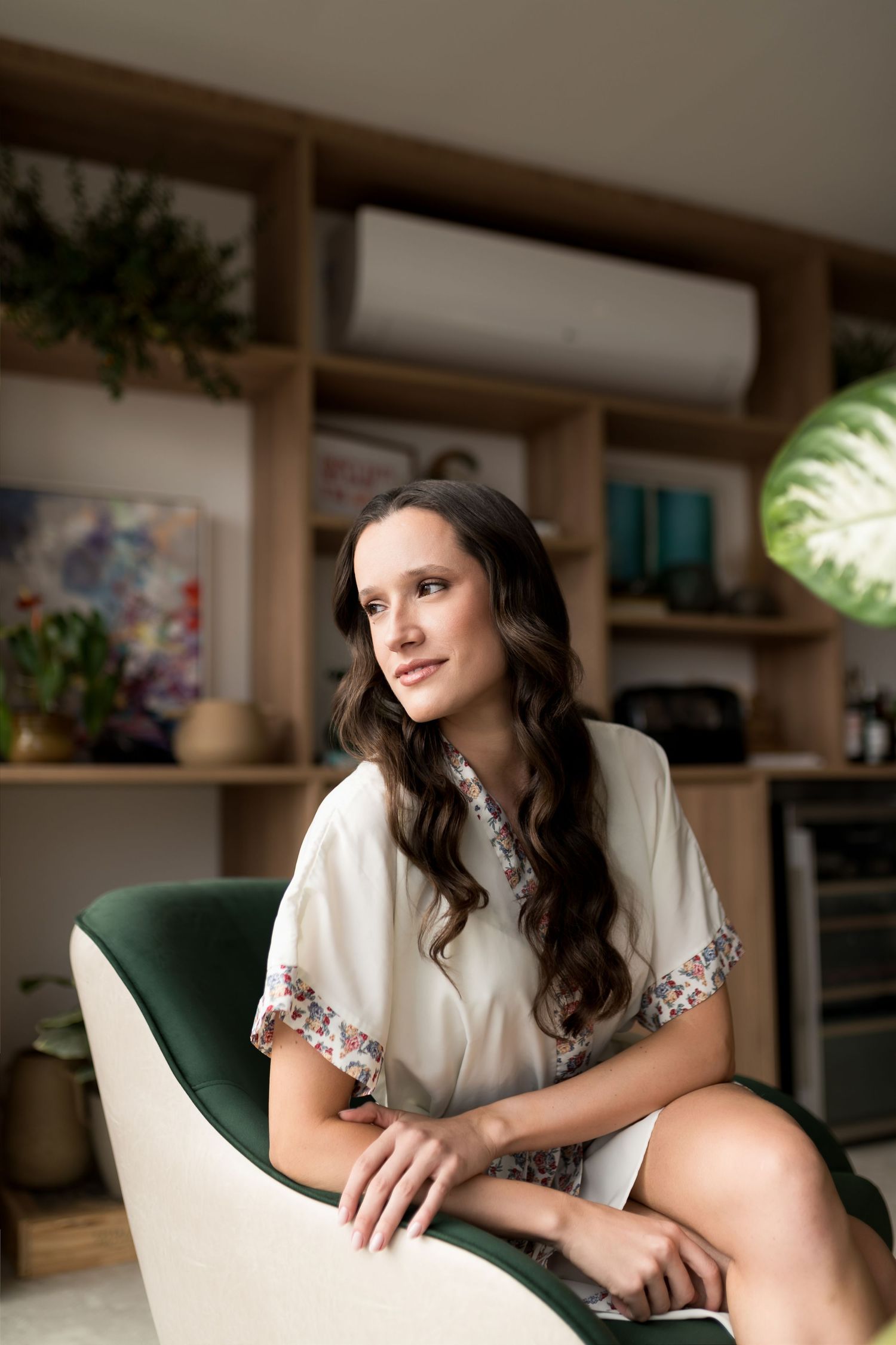 Young woman with long dark hair sits in a green velvet chair, wearing a floral blouse, gazing thoughtfully in a modern room.