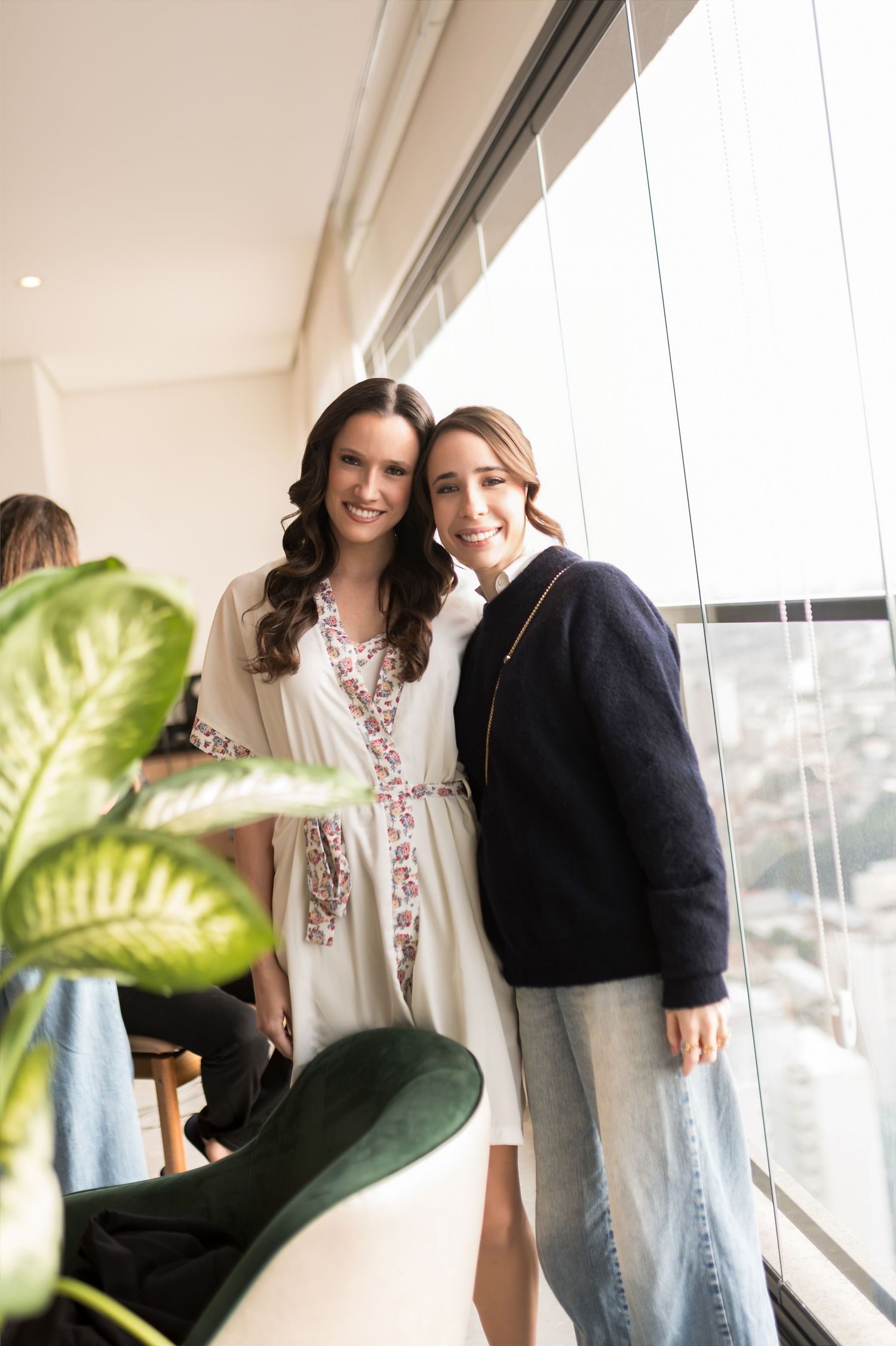 Two smiling women posing together near large windows in a bright, modern interior space with a plant in foreground.