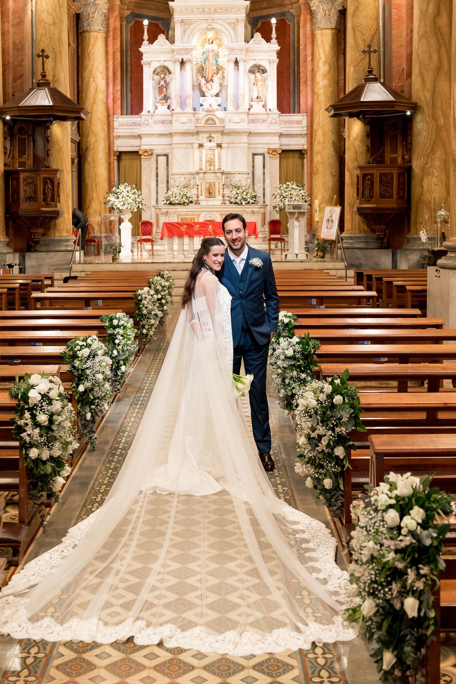 Bride and groom pose at church altar after wedding ceremony, bride wearing long lace veil trailing down aisle.