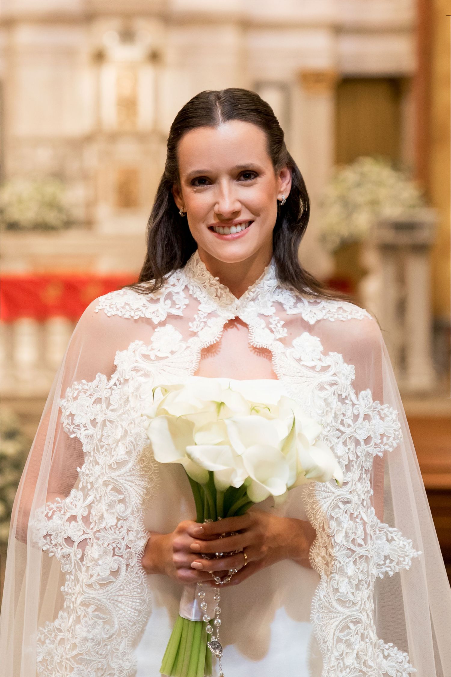 Smiling bride in white lace wedding dress holding bouquet of white calla lilies inside a church.