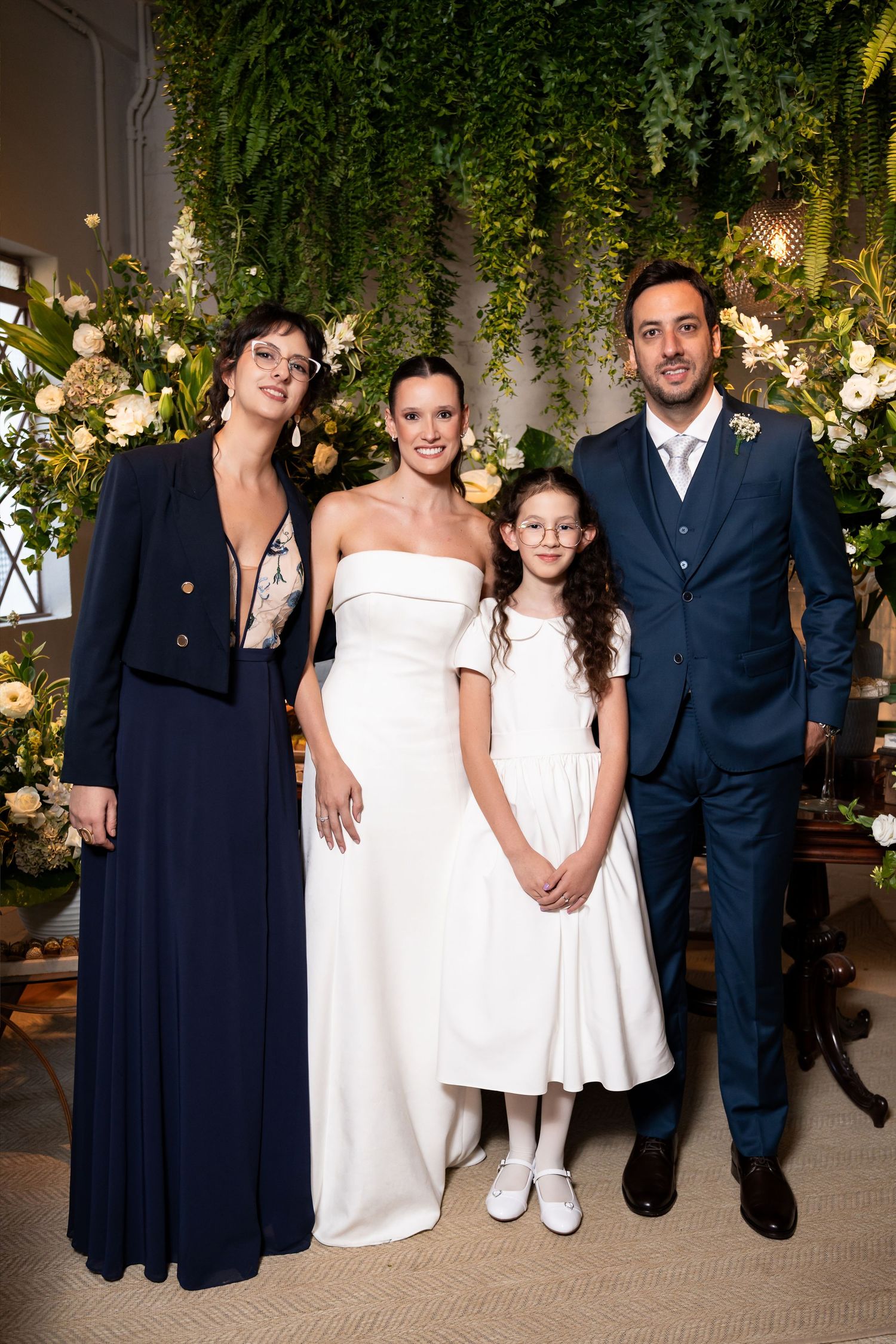 Wedding party poses with bride in white gown, groom in navy suit, flower girl, and guest before floral backdrop.