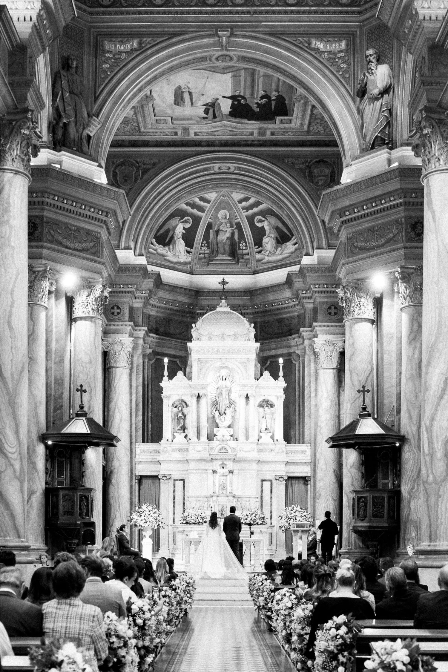 Black and white photo of a wedding ceremony inside an ornate Catholic church with grand altar and seated guests.