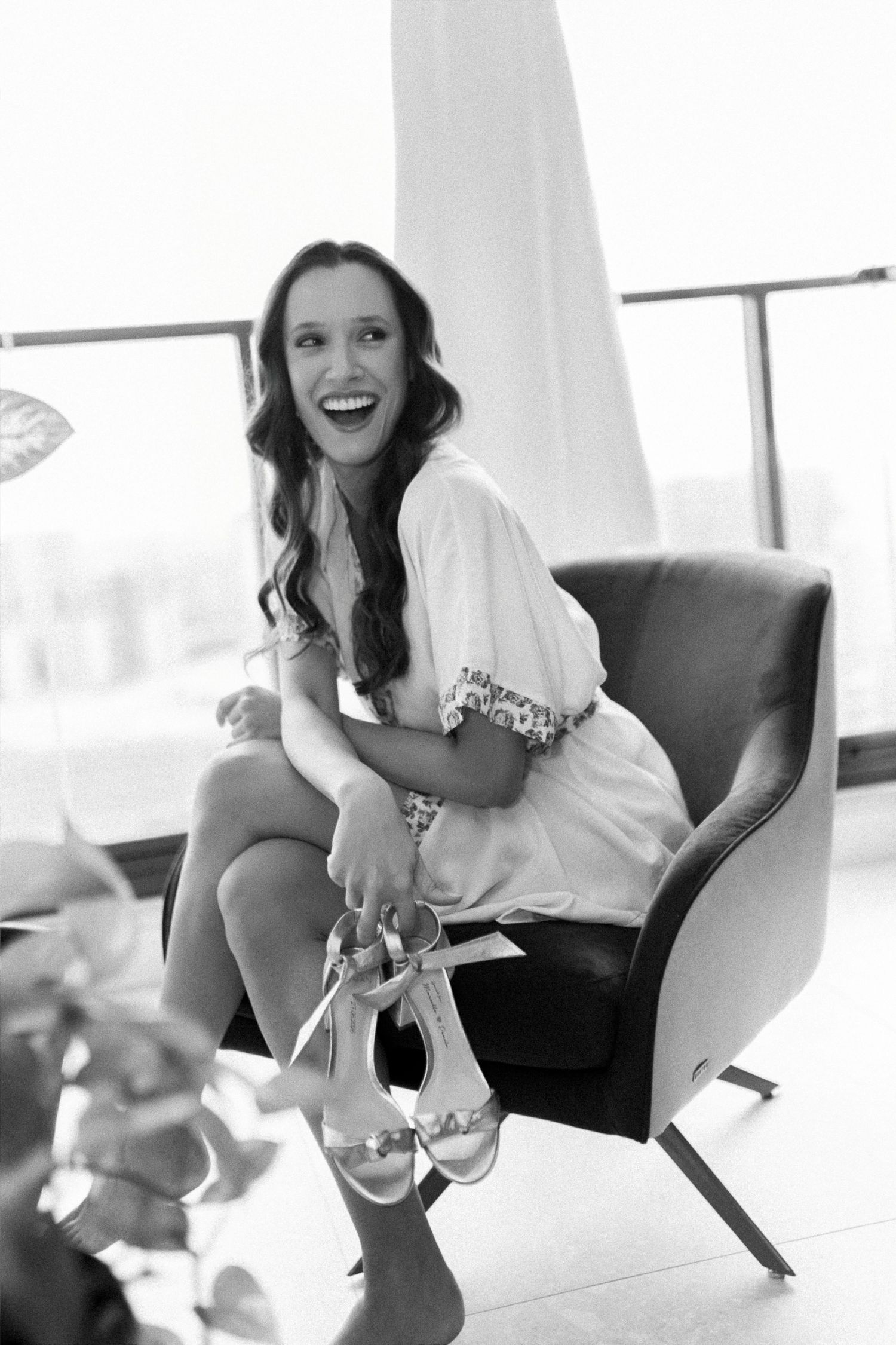 Black and white photo of a laughing woman in a white dress sitting in a chair holding heeled sandals.
