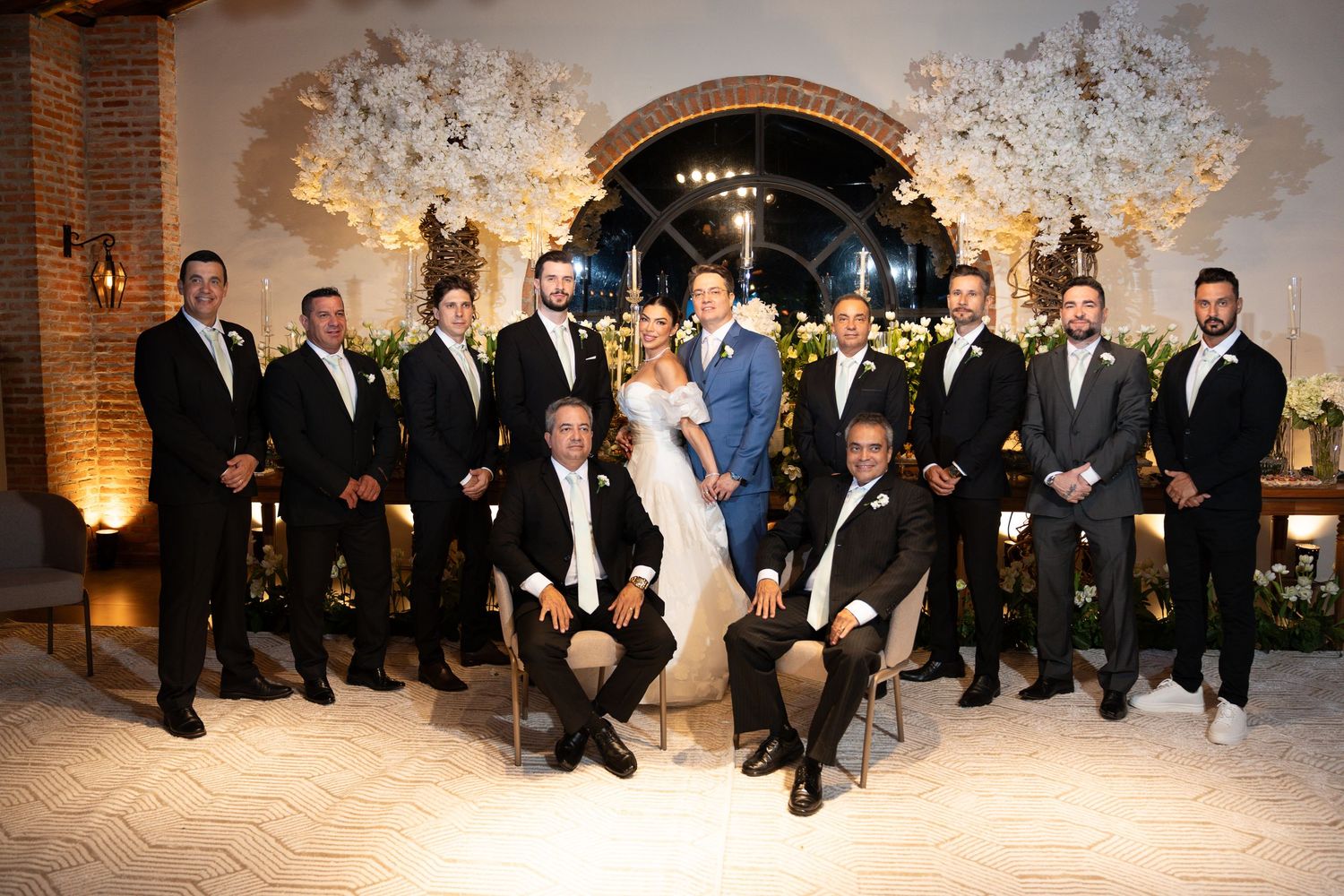 Wedding party poses elegantly in a floral-decorated venue, bride and groom centered among groomsmen in black tuxedos.