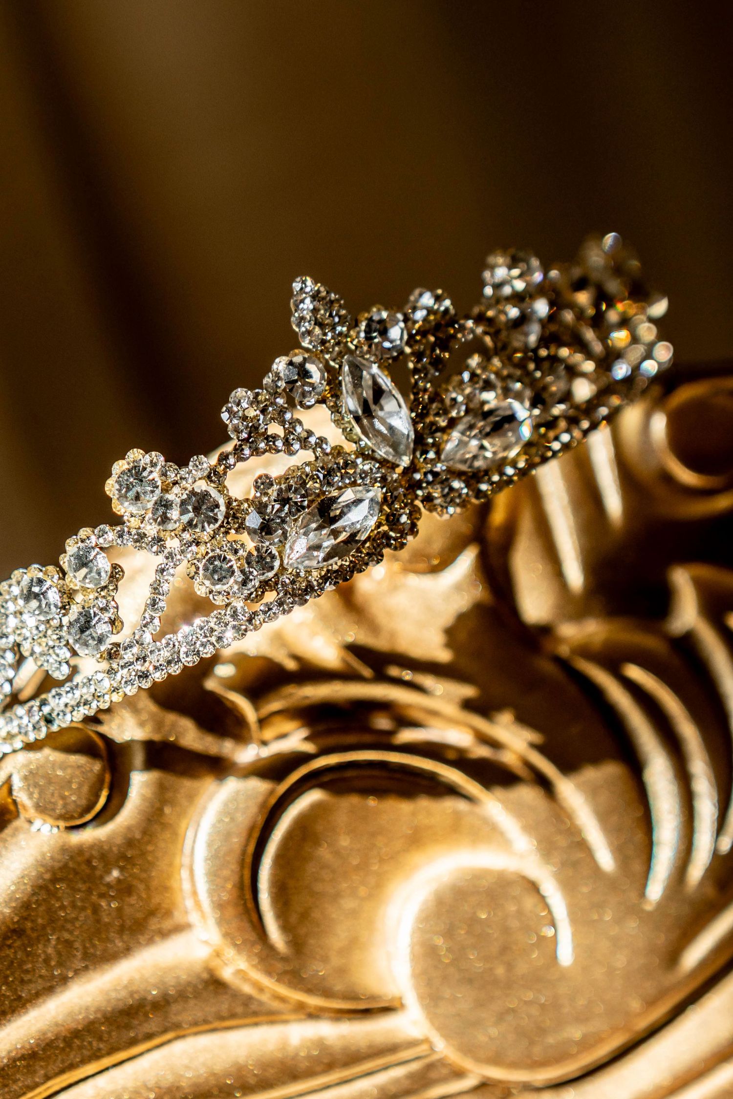 Elegant diamond tiara with sparkling crystal gems resting on a golden ornate surface with decorative swirl patterns.