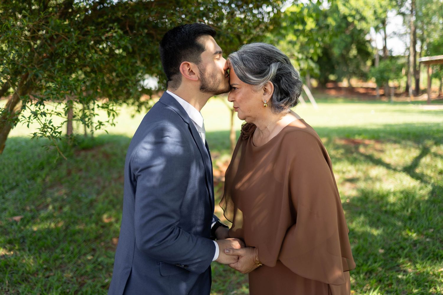 A young man in a suit kisses an older woman on the cheek while holding hands in a lush green park.