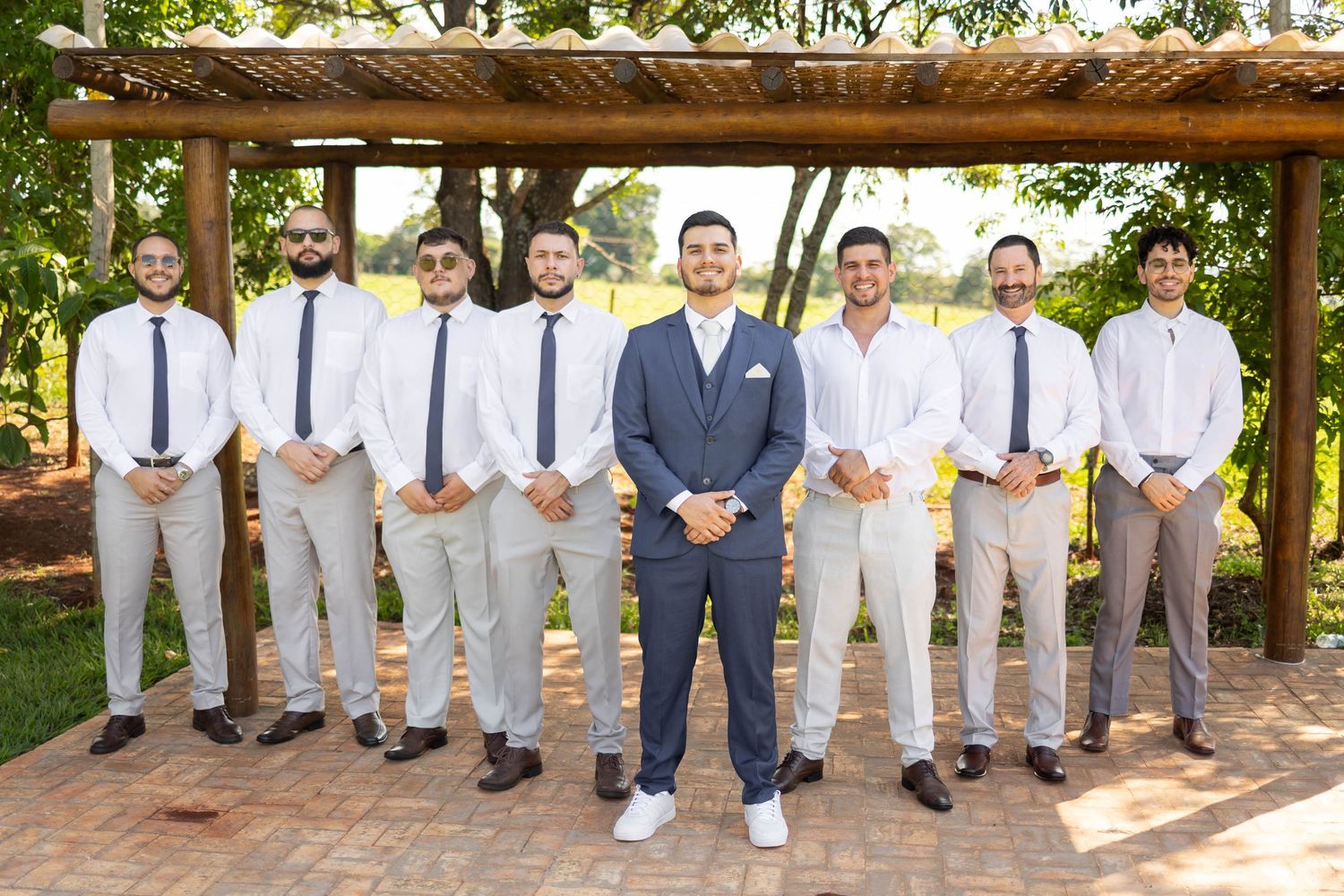 Groom in navy suit poses with groomsmen in white shirts and khaki pants outdoors under a wooden pergola.