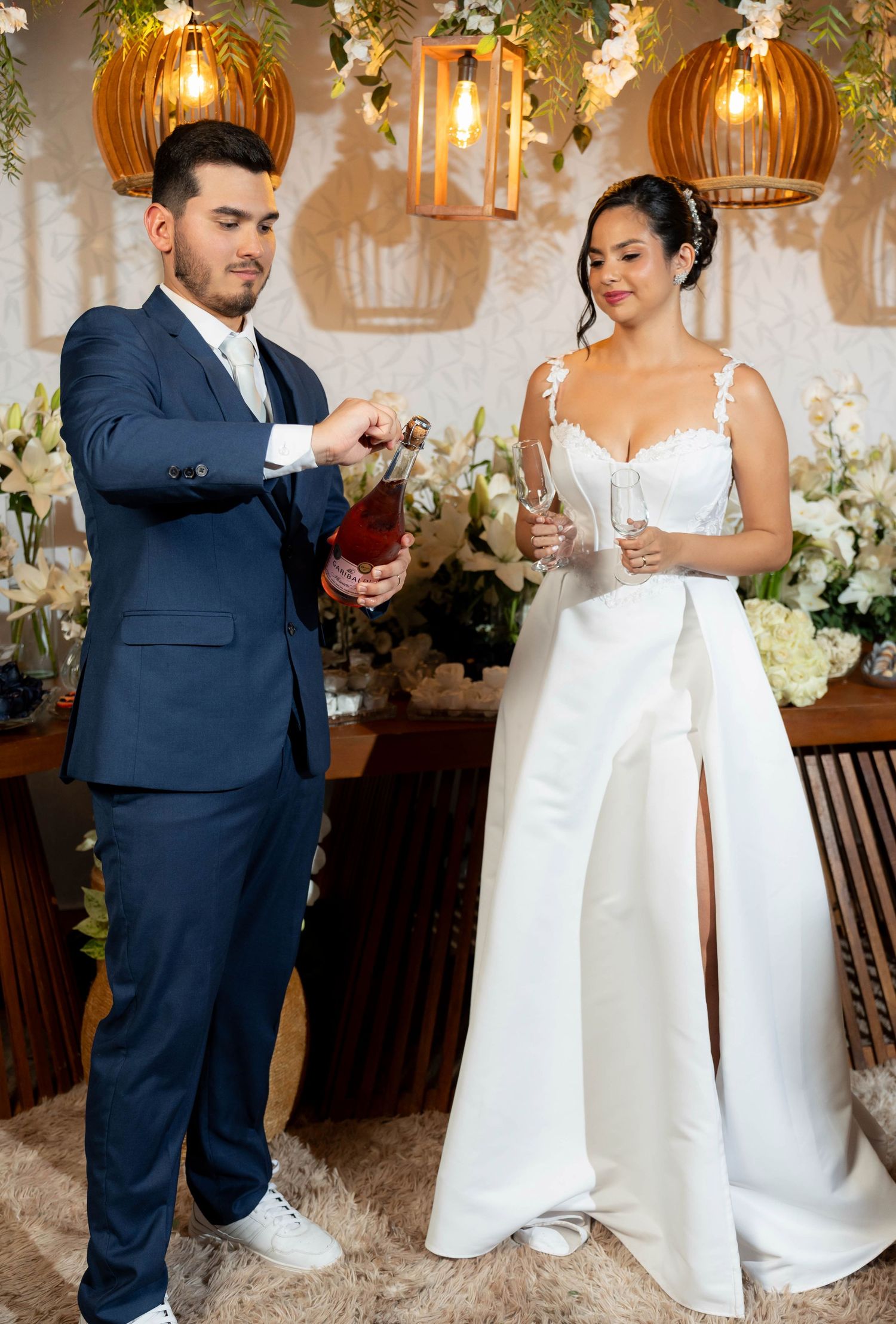 A groom in a navy suit pours champagne for his bride in a white wedding jumpsuit at their elegant floral reception.