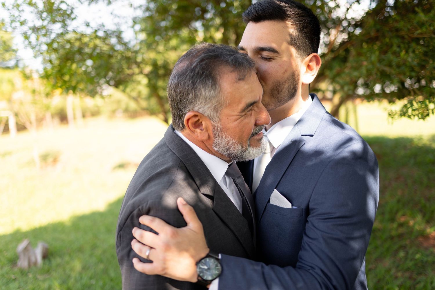 Two men in suits share an emotional embrace outdoors, with the younger man kissing the older man on the cheek.