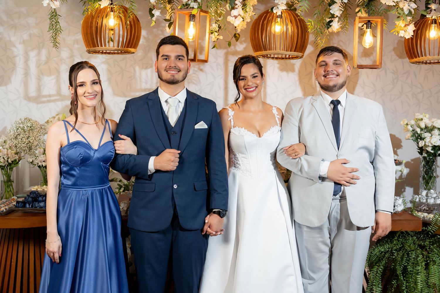 Wedding party of four posing indoors, two in blue formal wear and bride in white gown with groom in gray suit.