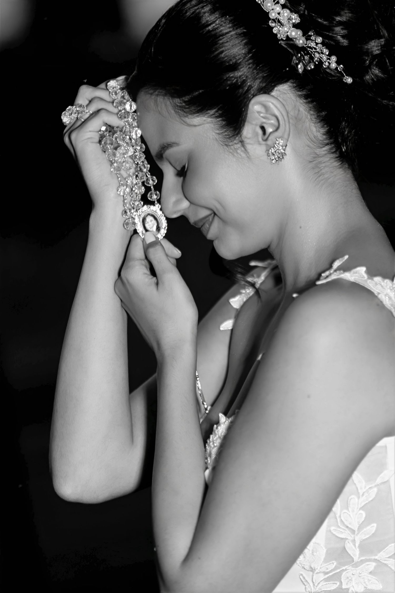 Black and white photo of a bride in a lace gown holding rosary beads to her forehead with eyes closed, smiling softly.