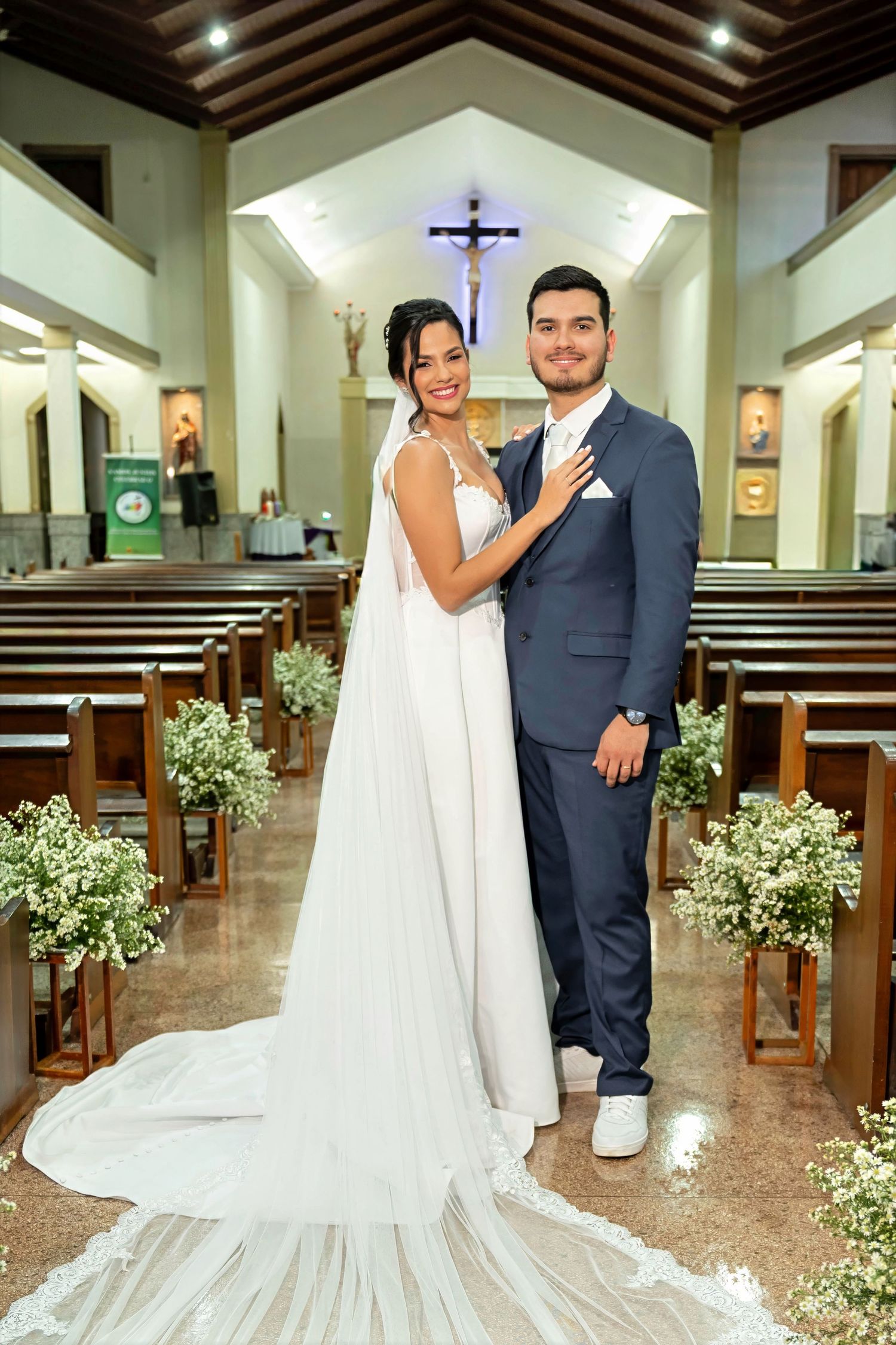 Bride in white gown with long veil and groom in navy suit pose together inside a decorated Catholic church.