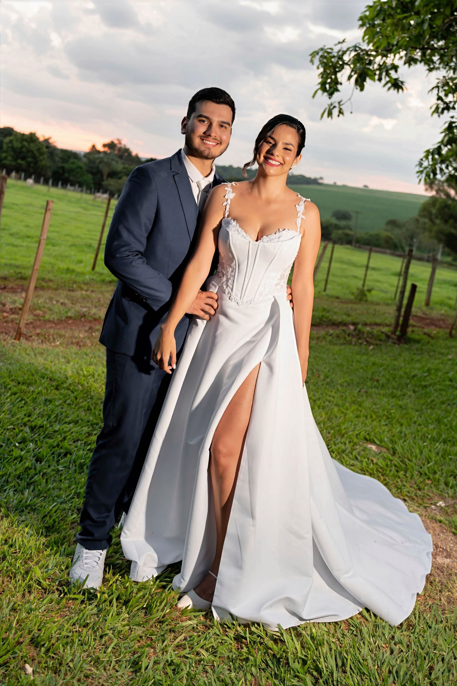 A smiling couple poses outdoors in wedding attire, groom in navy suit, bride in white gown with slit, green field backdrop.