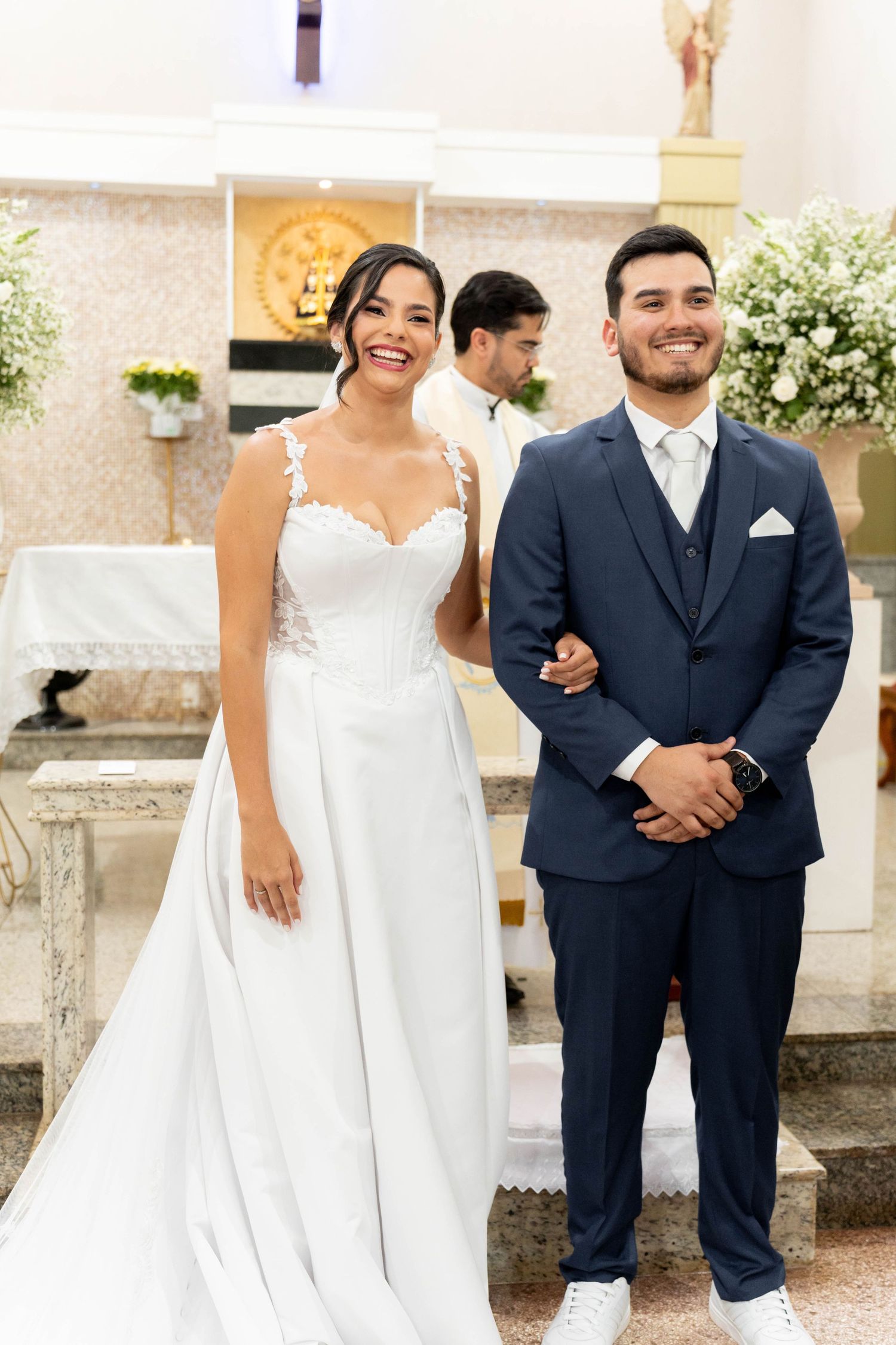 Smiling bride in white lace gown and groom in navy suit stand together at church altar during wedding ceremony.