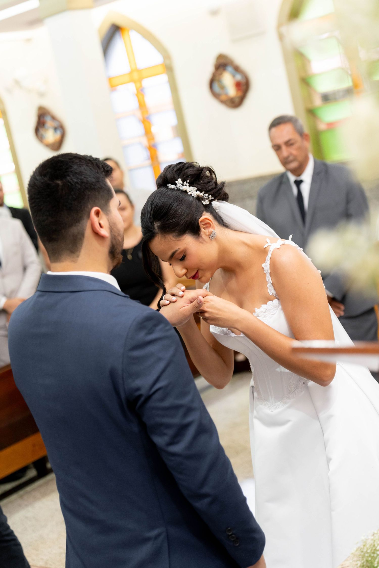 Bride in white gown kisses groom's hand during wedding ceremony in a church, with officiant standing in background.