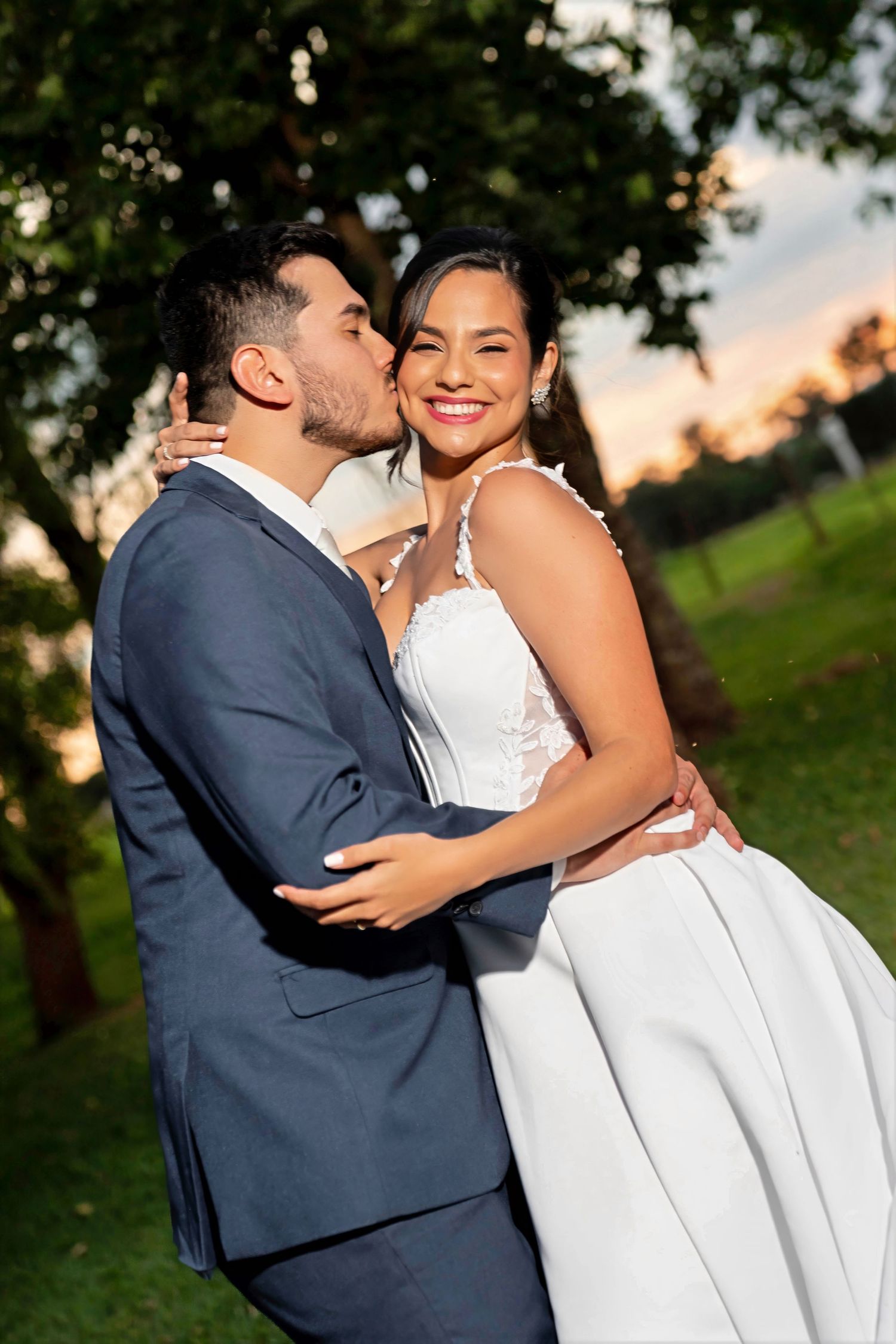 Happy bride in white wedding dress embraced by groom in navy suit sharing a romantic sunset kiss.