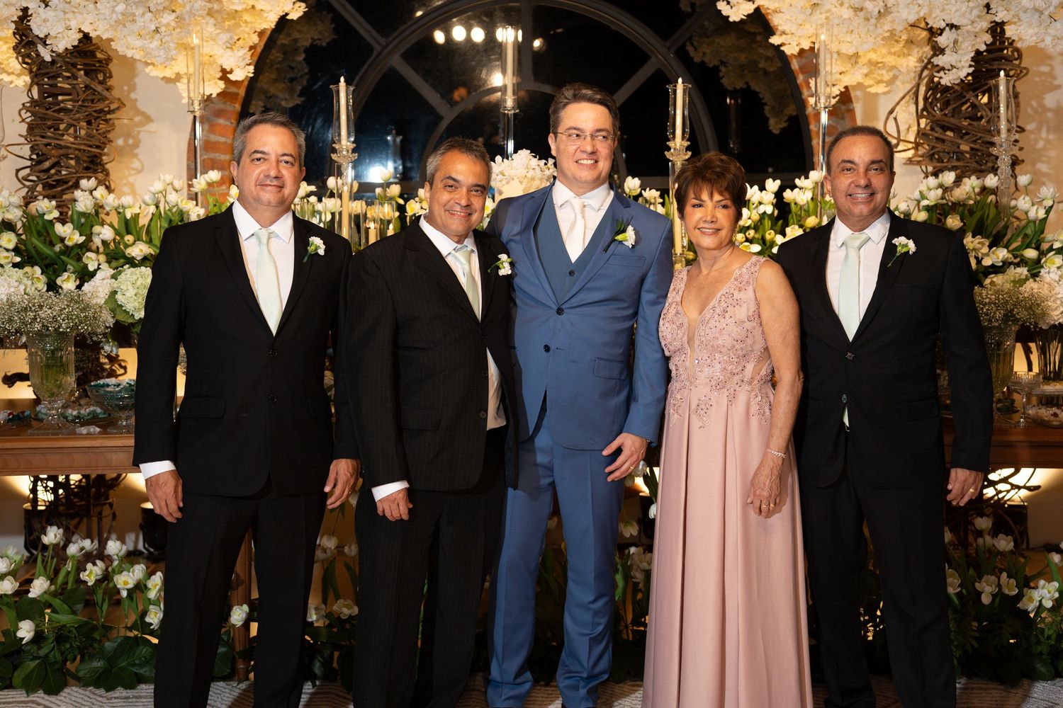 Five elegantly dressed guests pose together at a luxurious wedding reception with floral decorations.