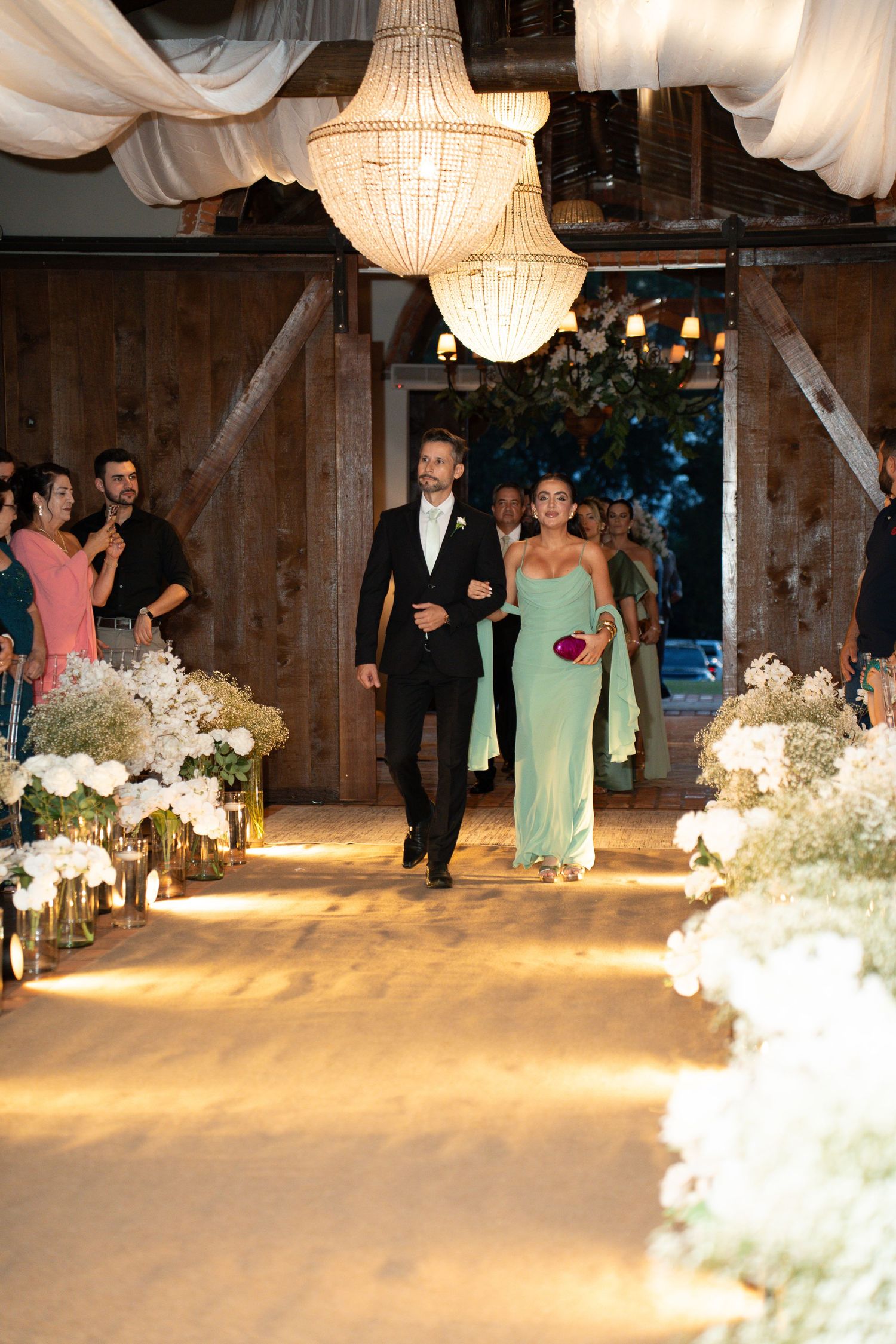 A couple walks down a floral-lined wedding aisle in a barn venue, man in tuxedo and woman in mint green gown.
