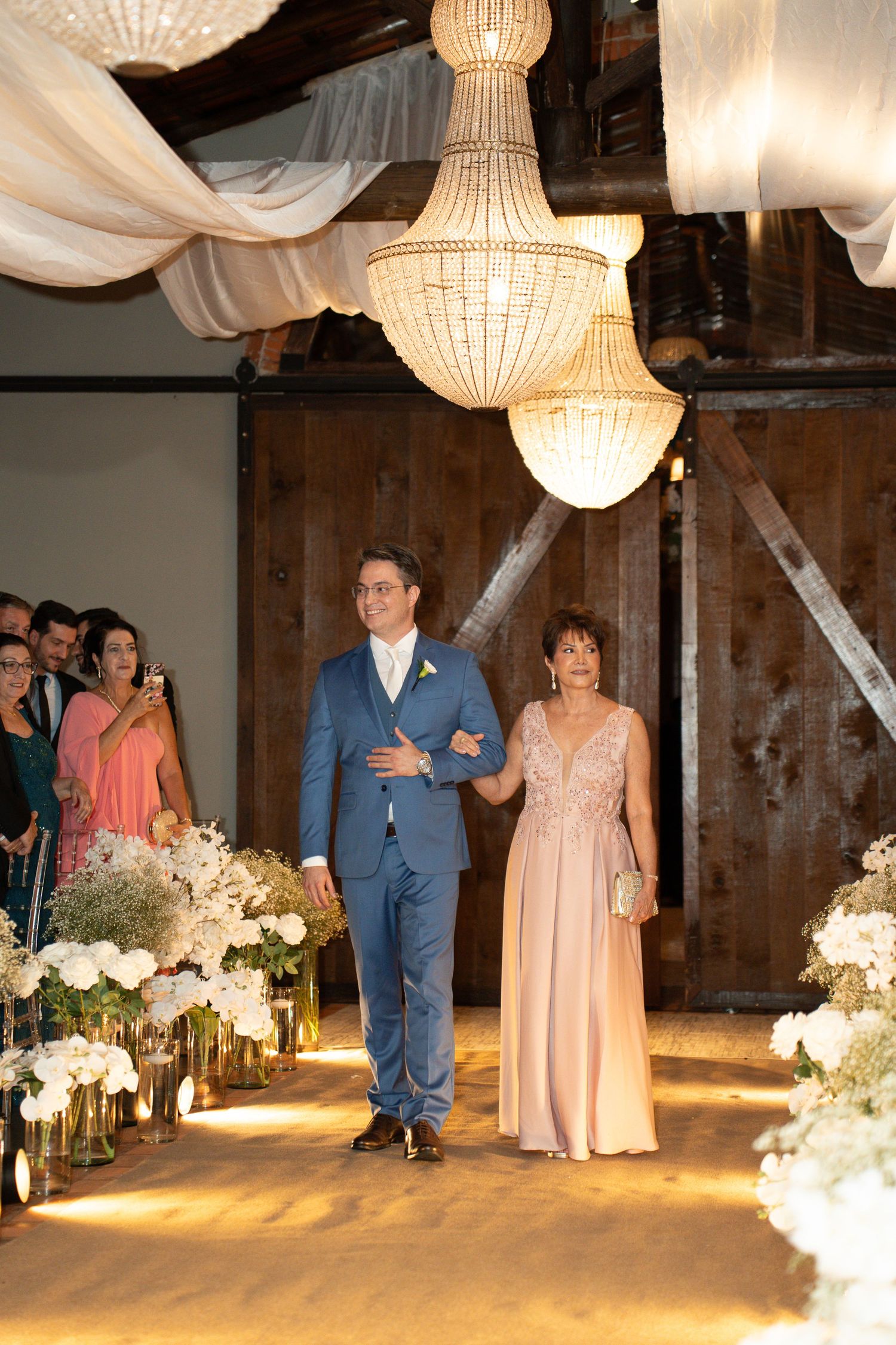 Groom in blue suit walks down wedding aisle with woman in pink gown under crystal chandeliers in rustic barn venue.