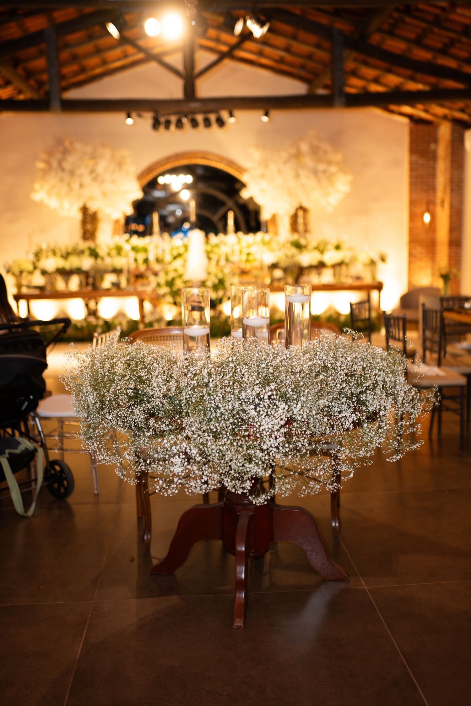 Elegant wedding reception with baby's breath floral centerpiece on wooden table, candlelit altar in rustic venue background.