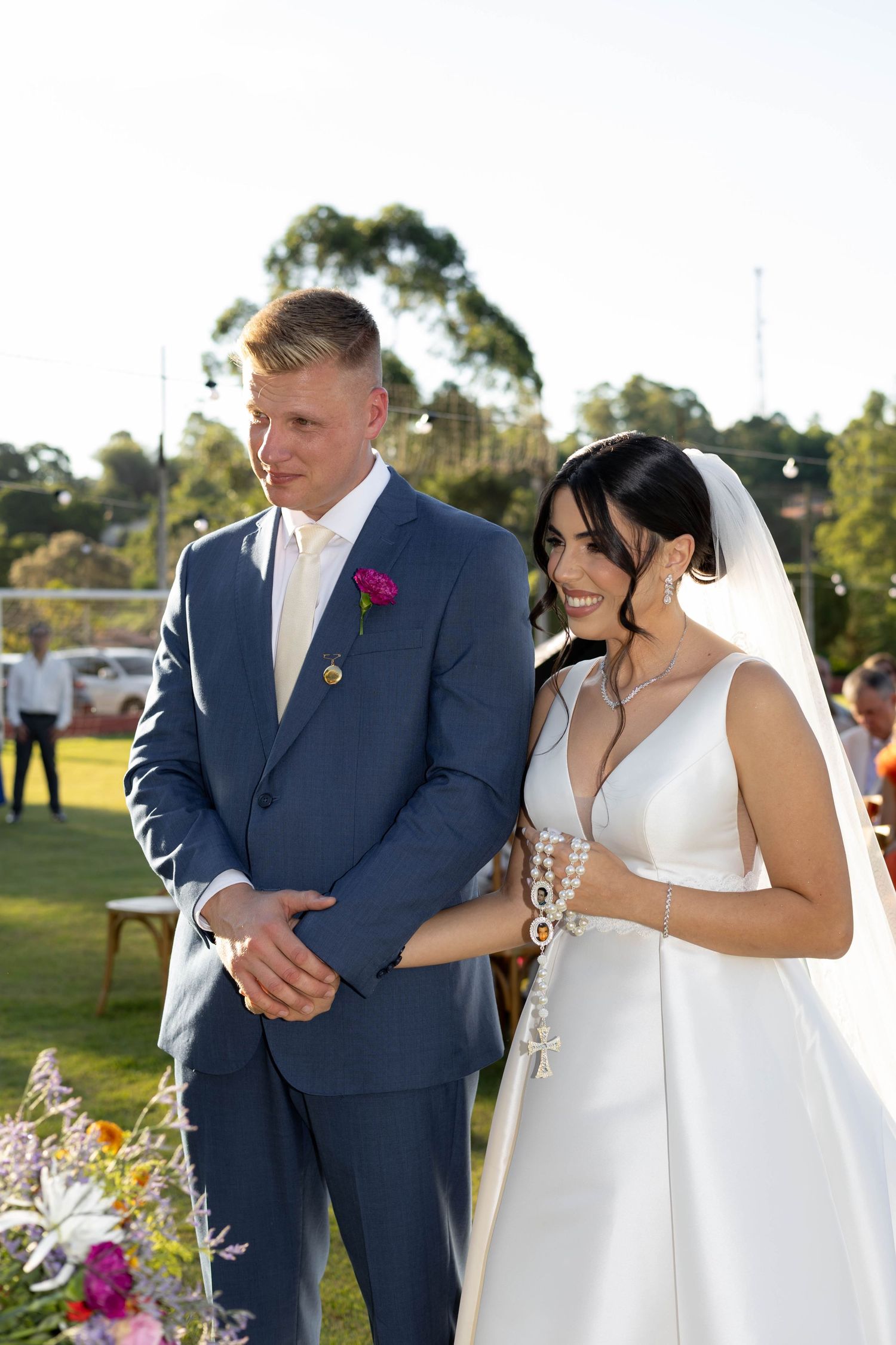 Bride in white gown and groom in blue suit smiling during outdoor wedding ceremony with green trees background.