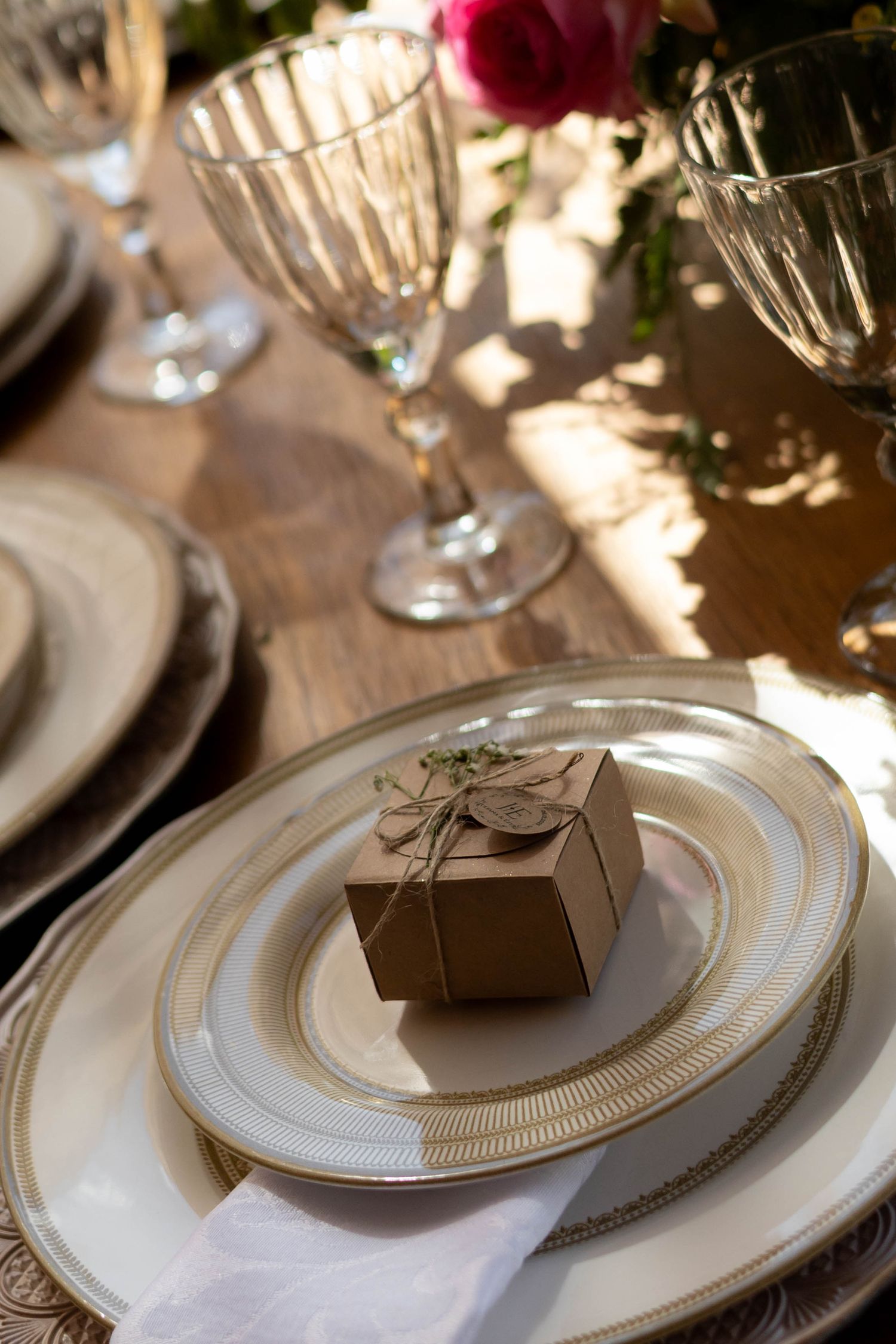 Elegant wedding table setting with crystal glasses, gold-rimmed plates, and a kraft paper gift box favor tied with twine.