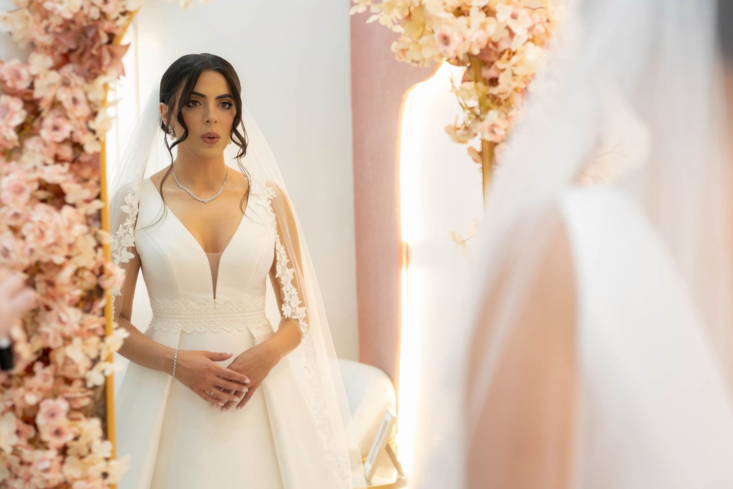 Bride in elegant deep V-neck white wedding gown with cape sleeves, standing before pink floral backdrop in bridal boutique.