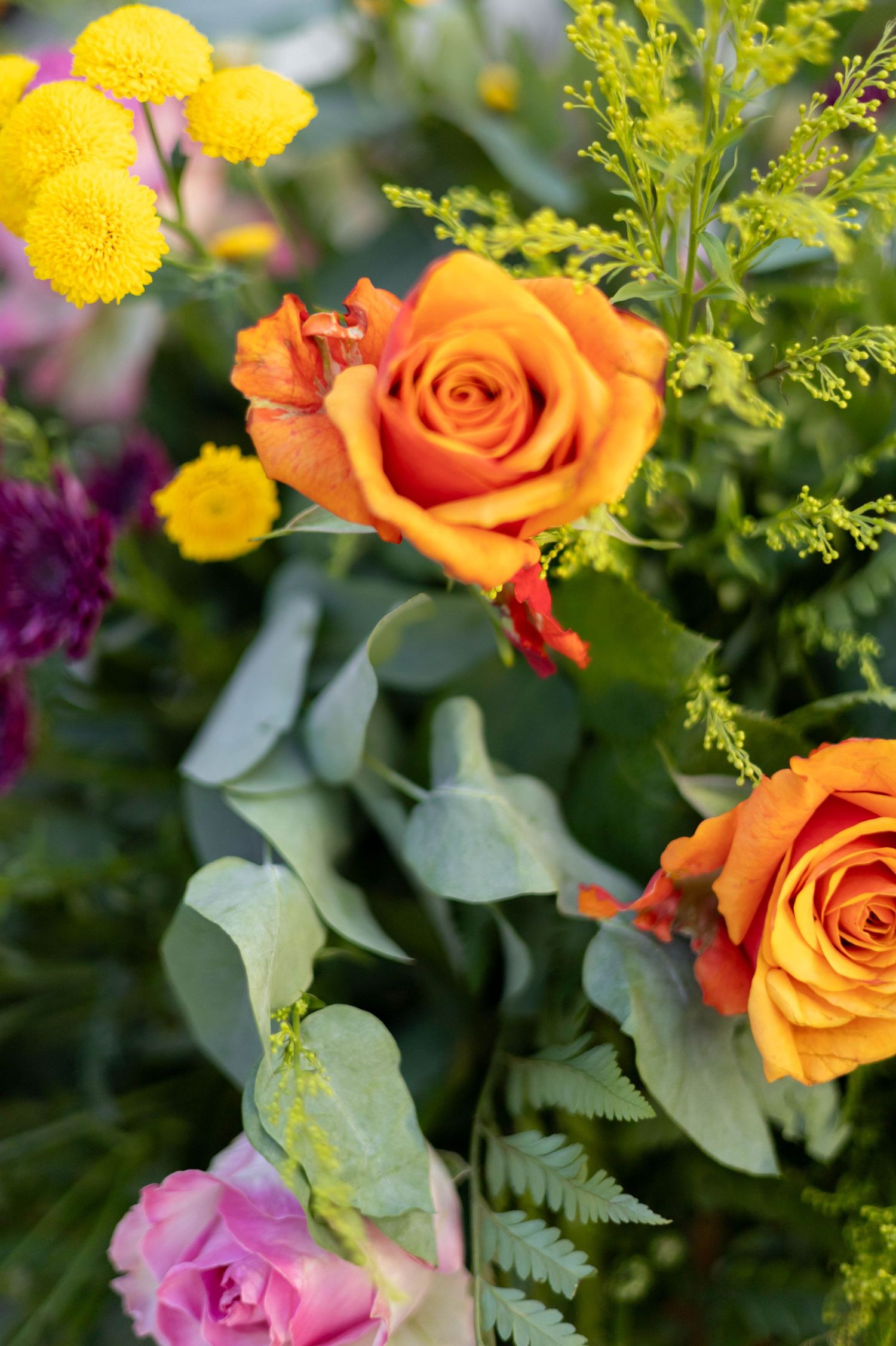 Vibrant bouquet featuring orange roses, yellow tansy flowers, and lush green foliage in a colorful floral arrangement.
