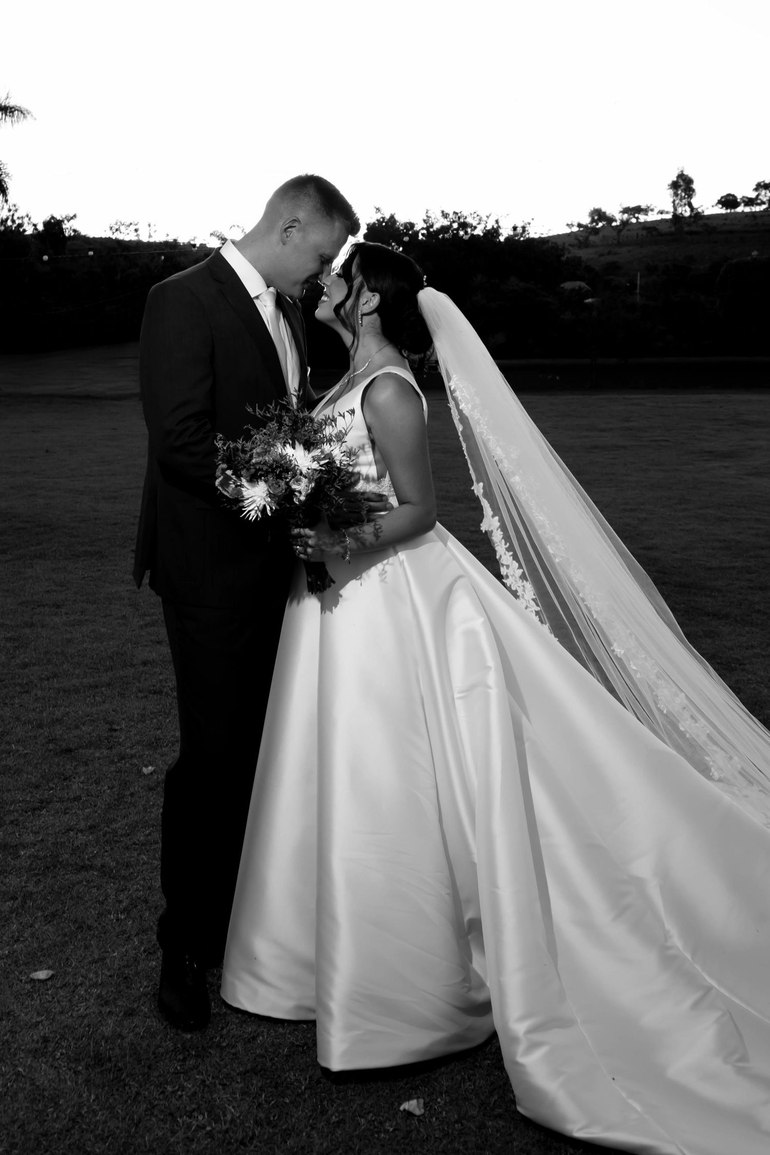 Black and white wedding photo of bride and groom sharing an intimate moment outdoors, bride holding bouquet with long veil.