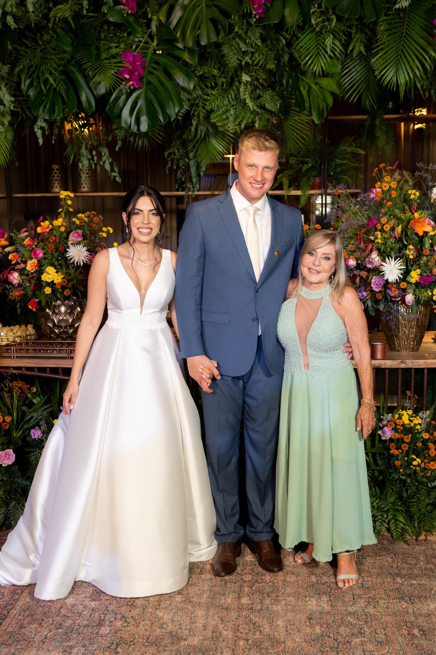 Bride in white gown and groom in blue suit pose with guest in mint dress at floral wedding backdrop.