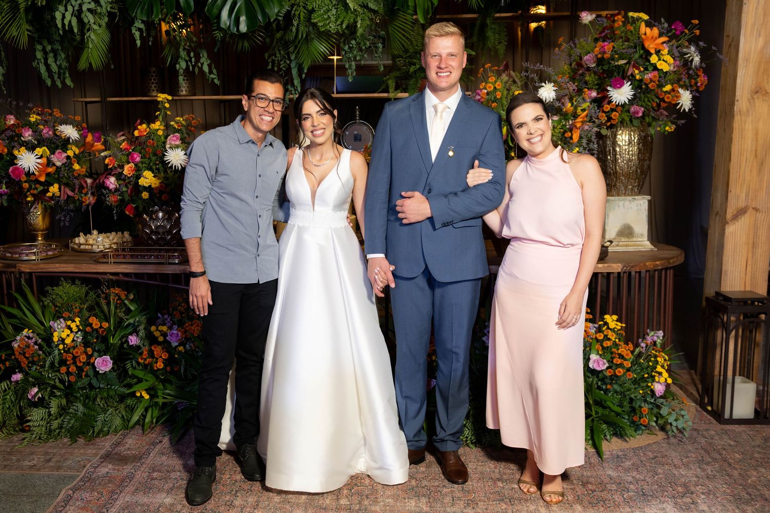 A bride in white gown and groom in blue suit pose with two guests amid lush floral arrangements at a wedding reception.
