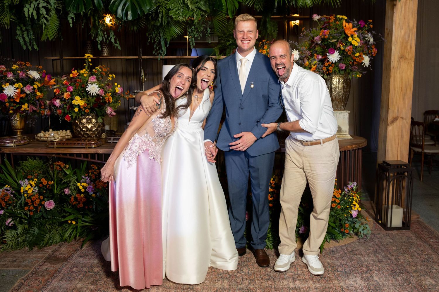 A joyful wedding group photo featuring a bride in white, groom in blue suit, and two guests posing among colorful floral decor.