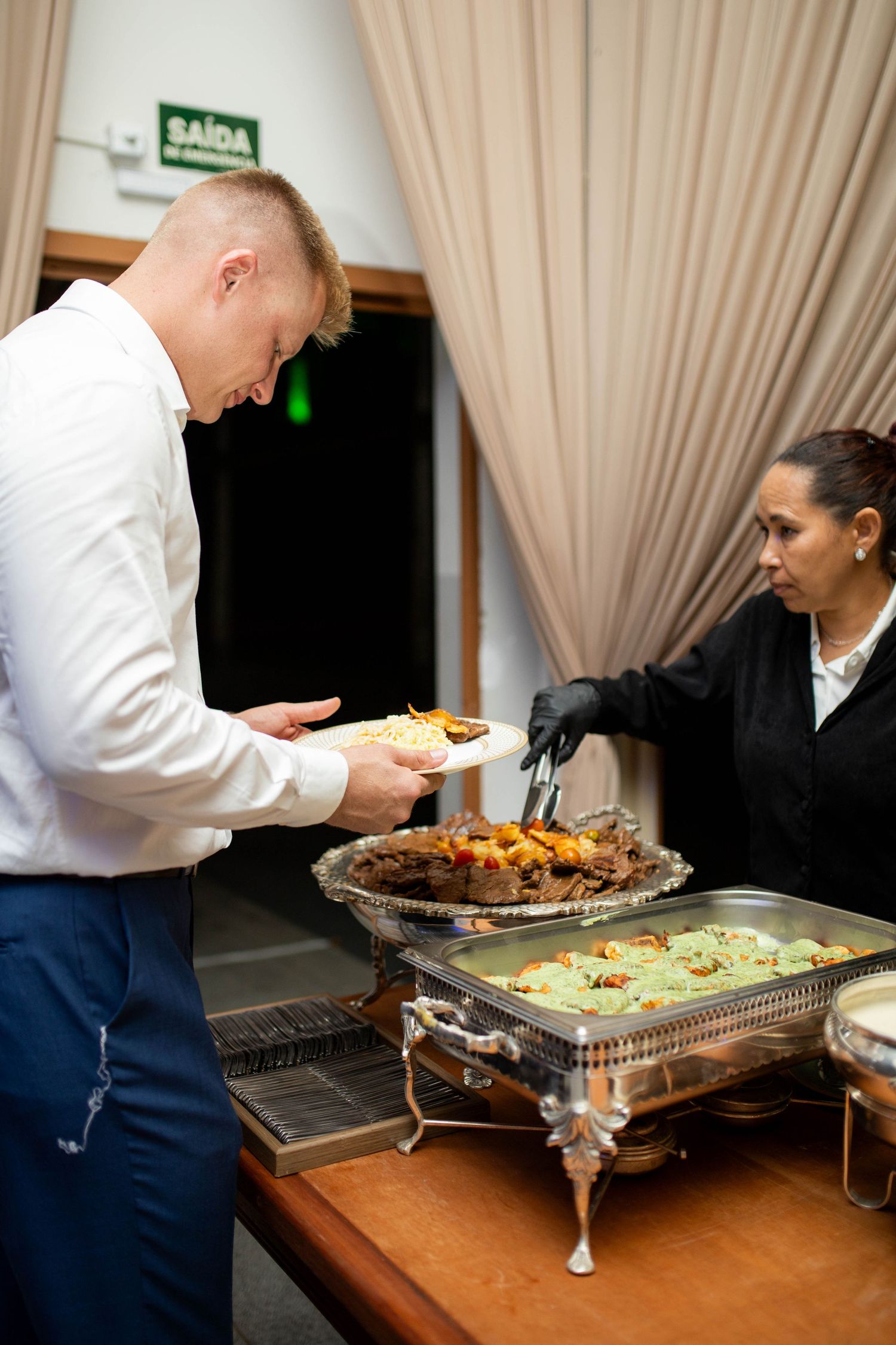 A server in black gloves serves food from a chafing dish to a man in a white shirt at a catered buffet event.