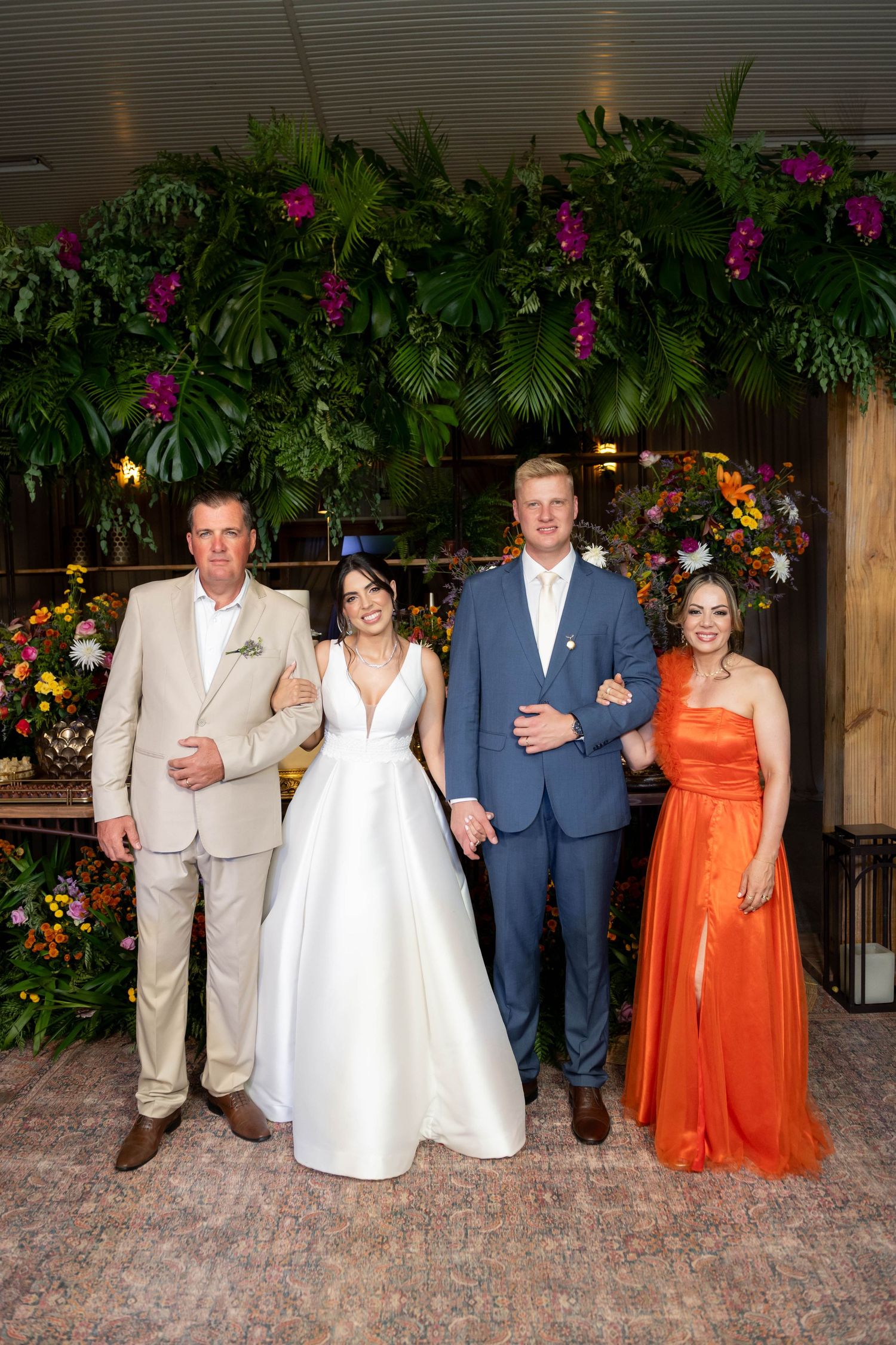 A bride in white gown and groom in blue suit pose with two guests before a lush tropical floral backdrop.