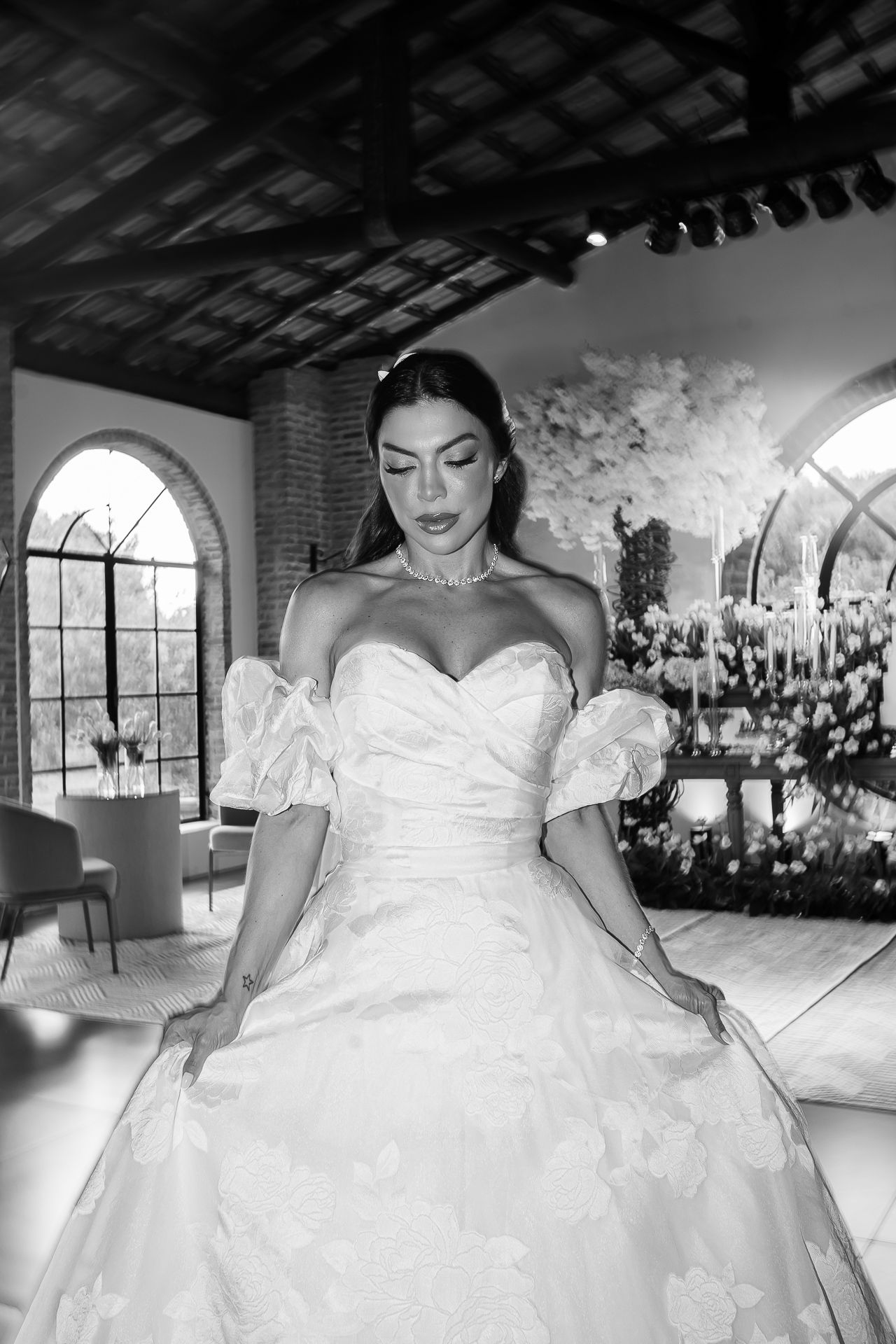 Bride in elegant off-shoulder ball gown wedding dress poses in black and white photo with floral backdrop.