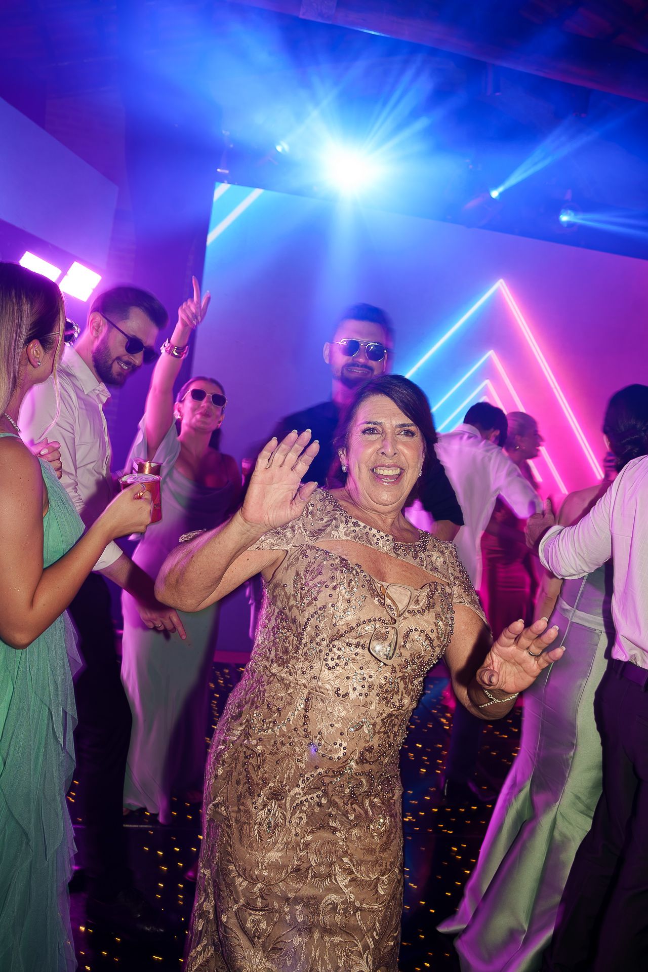 Woman in gold dress dancing joyfully at a vibrant party with colorful neon lights and guests in background.