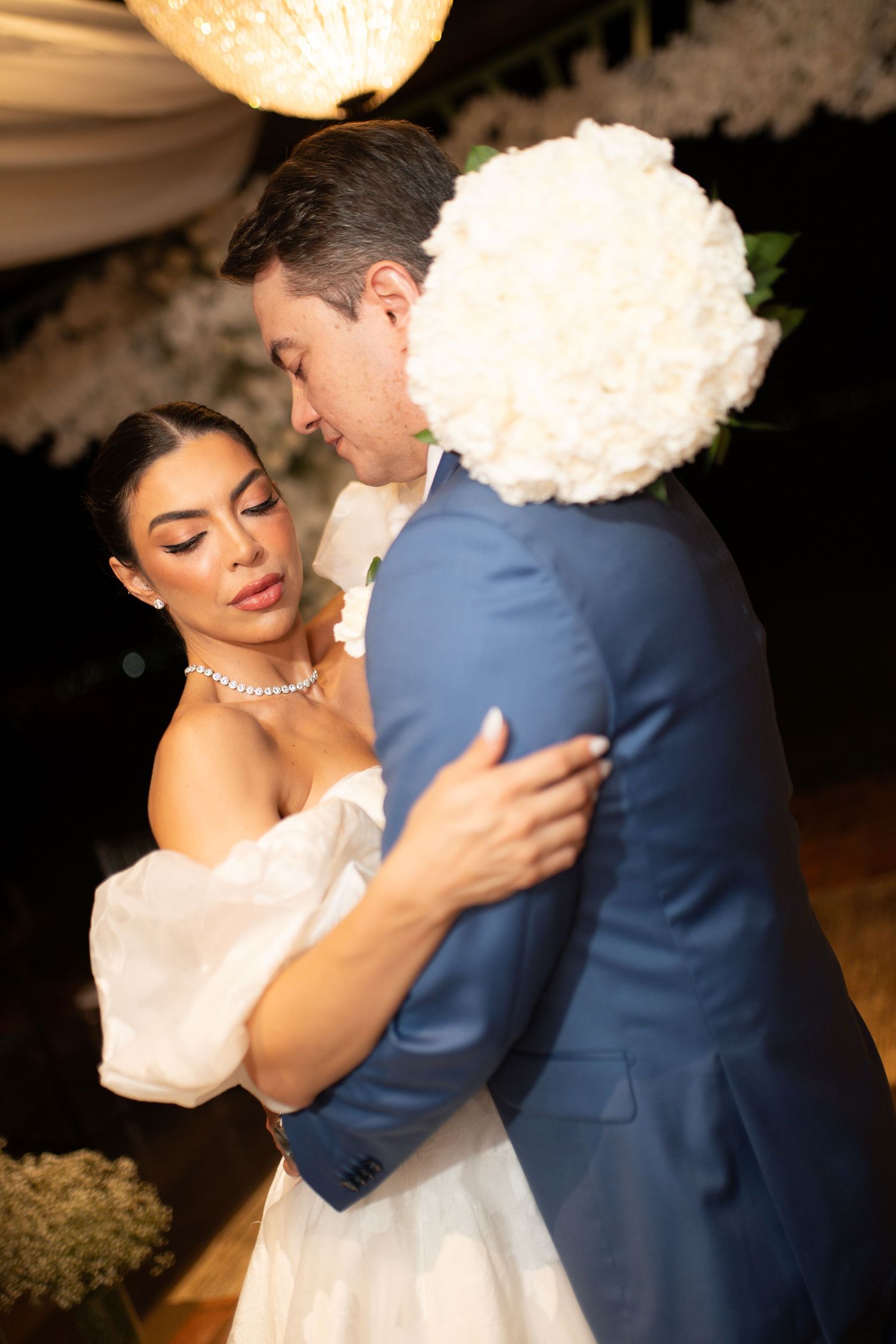 Bride and groom share first dance, bride in off-shoulder white gown holding rose bouquet, groom in blue suit.