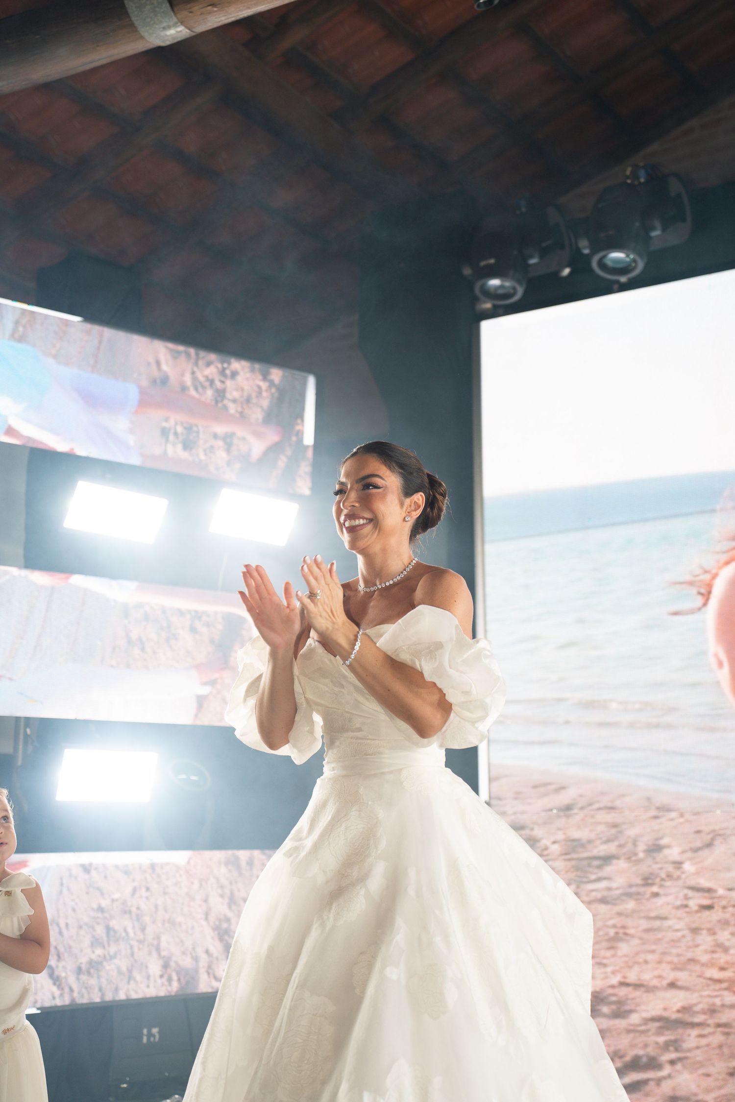 Smiling bride in white off-shoulder gown clapping joyfully at an elegant indoor wedding venue with bright lights.
