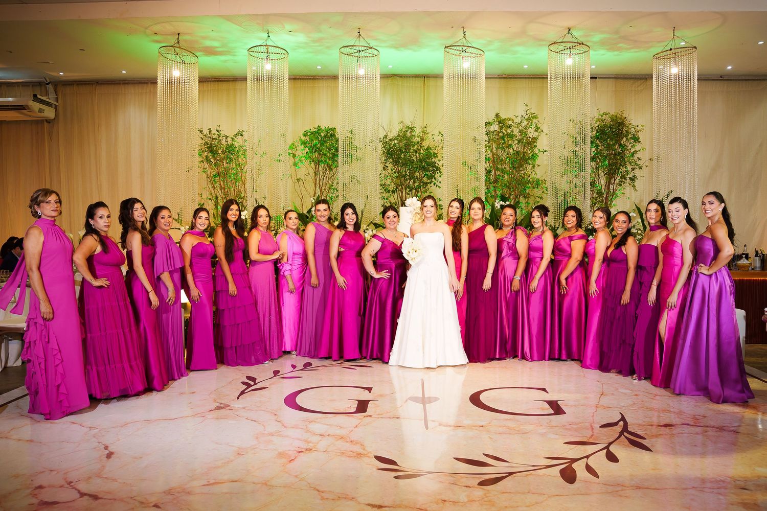 Bride in white gown poses with large group of bridesmaids in fuchsia dresses on monogrammed dance floor.