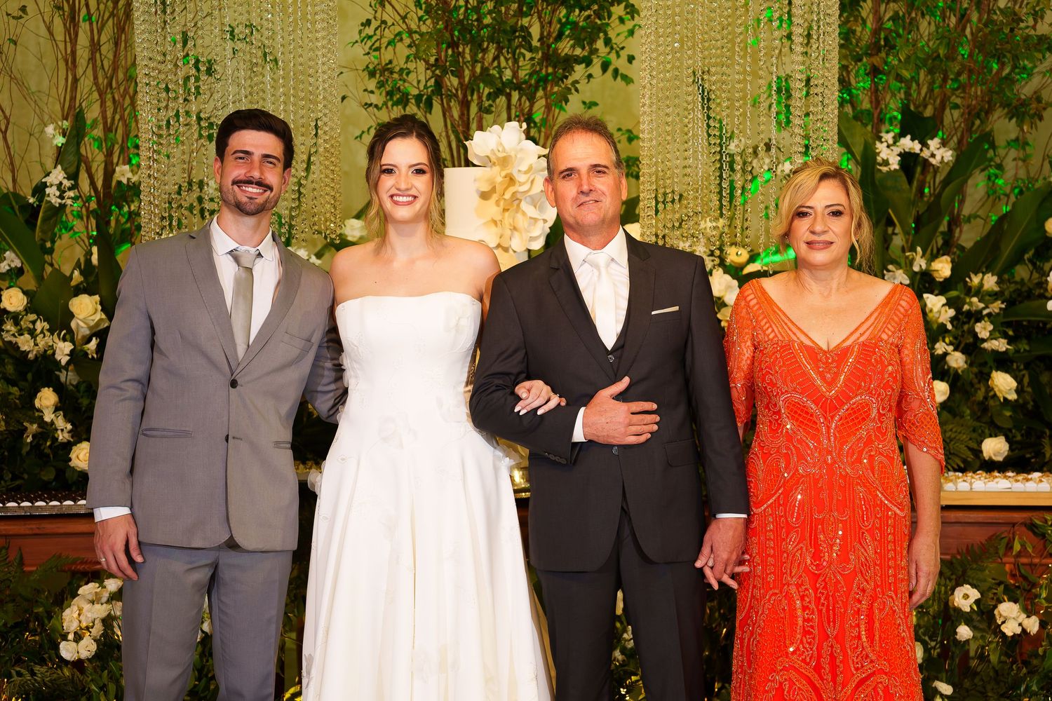 Wedding party poses together, bride in white gown flanked by groom in dark suit and guests in formal attire.