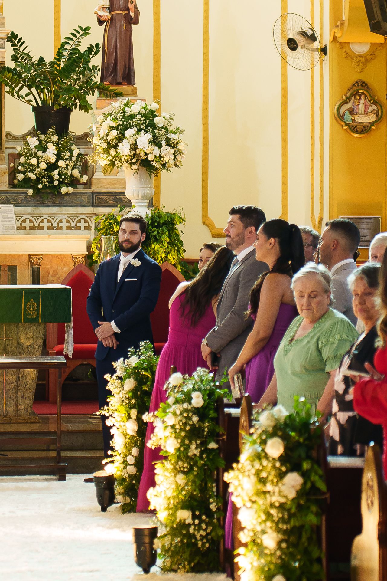 Groom in navy suit sits at church altar during wedding ceremony with guests and floral arrangements.