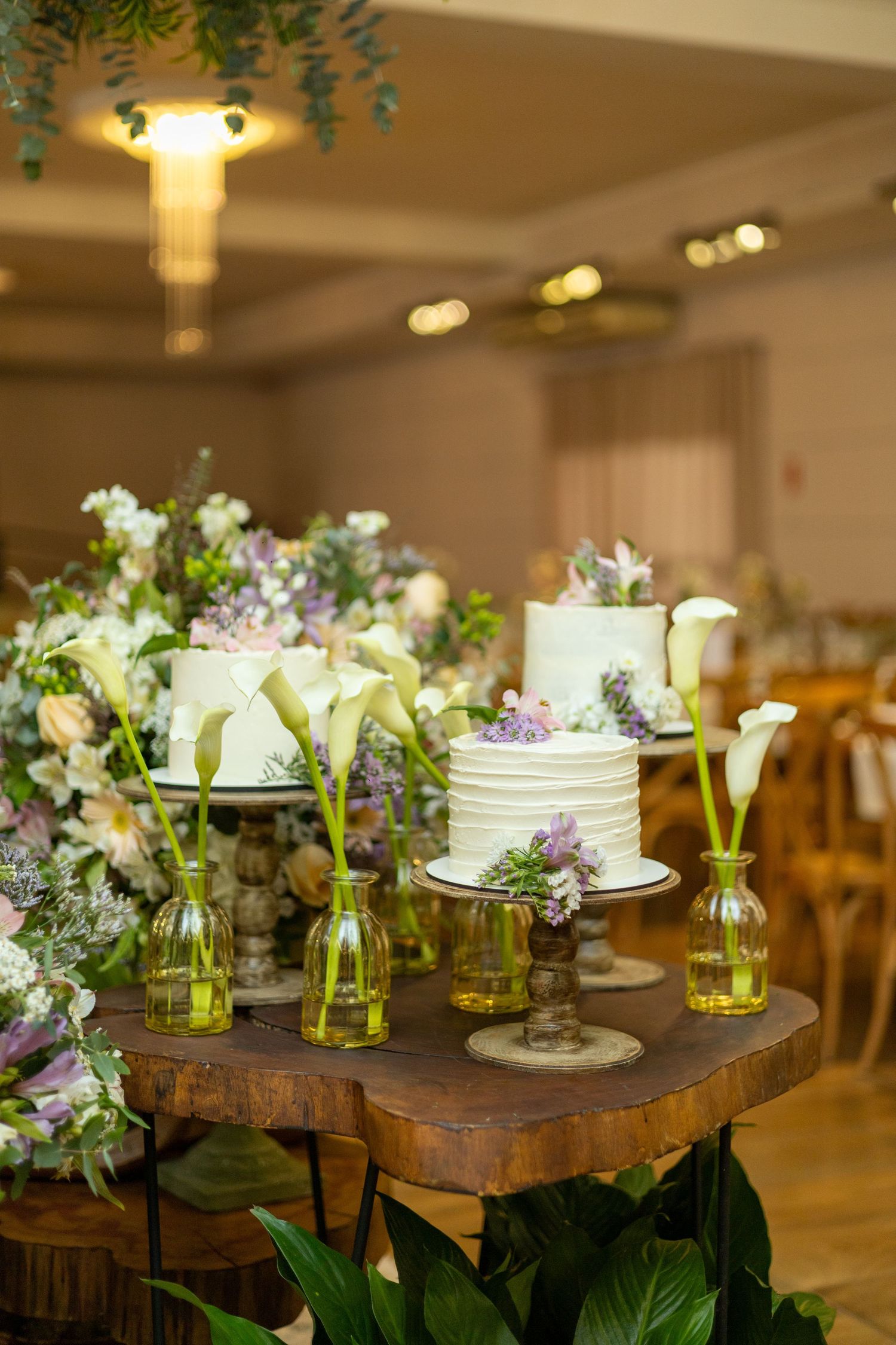 Elegant wedding cake table with white cakes, calla lilies in green vases, and lush floral arrangements on a rustic wooden stand.