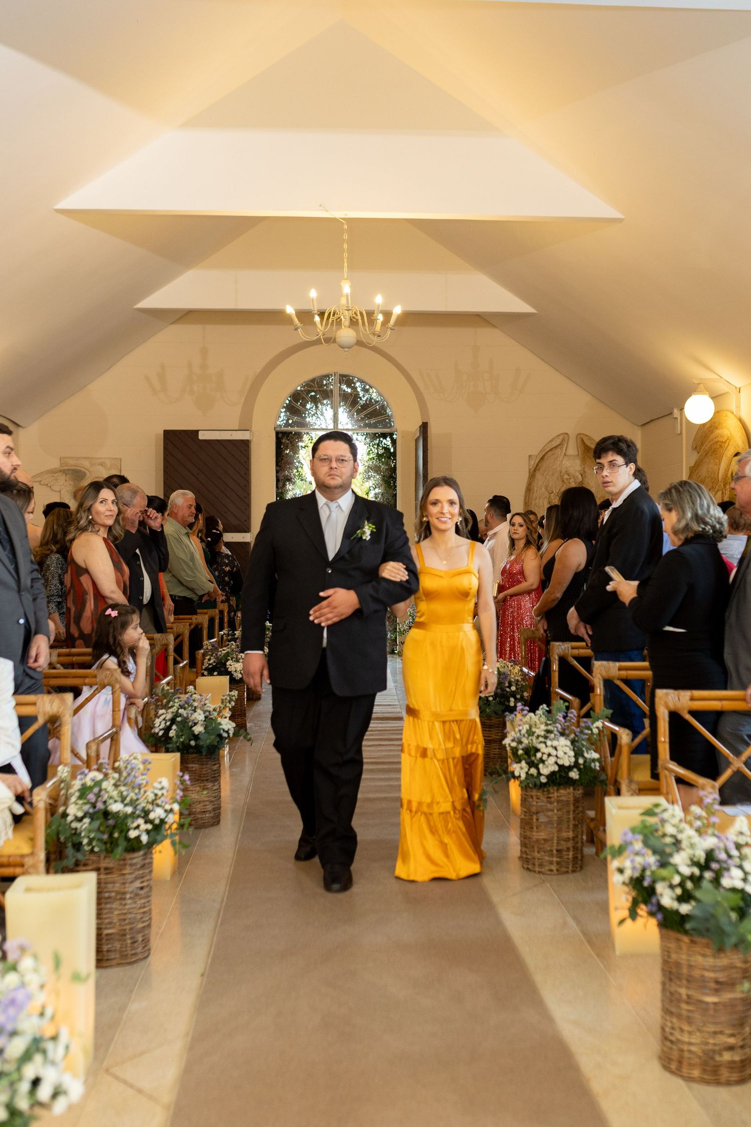 A couple walks down a chapel aisle, man in black suit, woman in yellow gown, flanked by floral arrangements.