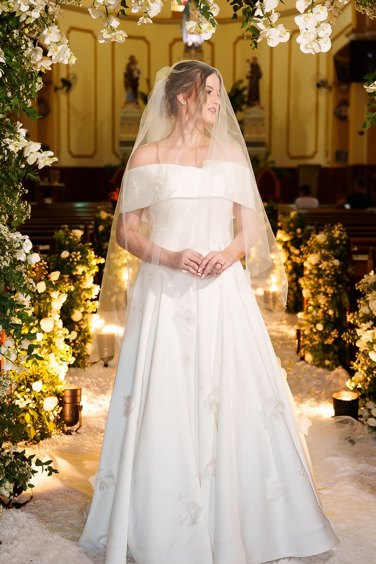 Bride in off-shoulder white ball gown with veil stands in flower-adorned church aisle with candles.