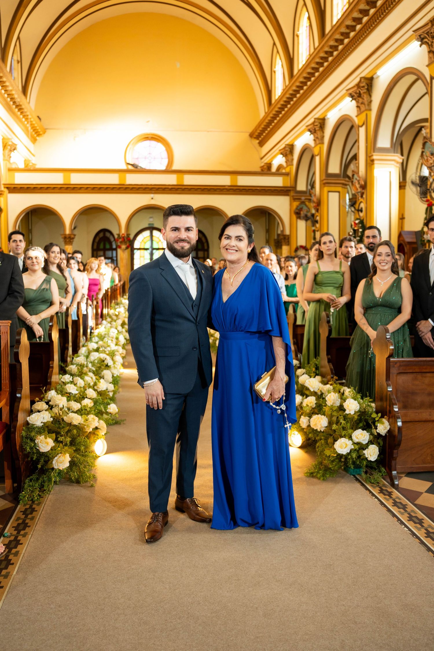 A man in a navy suit and woman in a royal blue gown pose together in an elegant church aisle at a wedding.