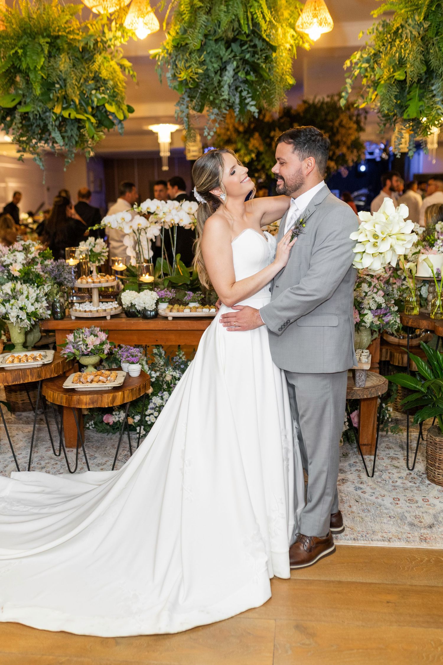 Bride in white gown and groom in gray suit embrace at elegant wedding reception with floral decor and greenery.