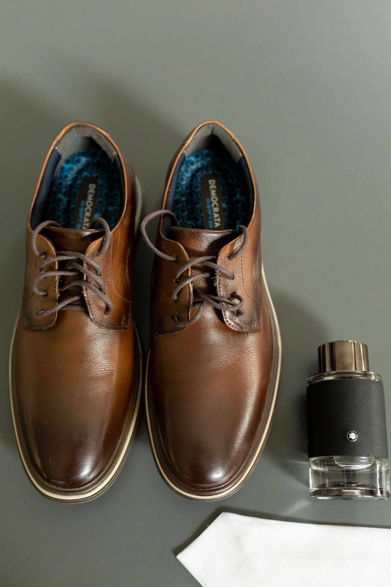 Brown leather lace-up dress shoes displayed beside a Mont Blanc cologne bottle on a gray surface.